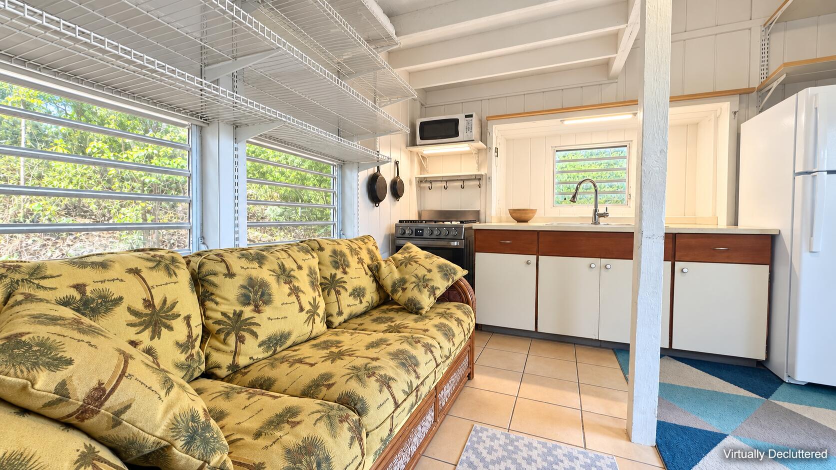 Open-plan living area and kitchen with a palm-print sofa, white cabinetry, and wide windows facing greenery.