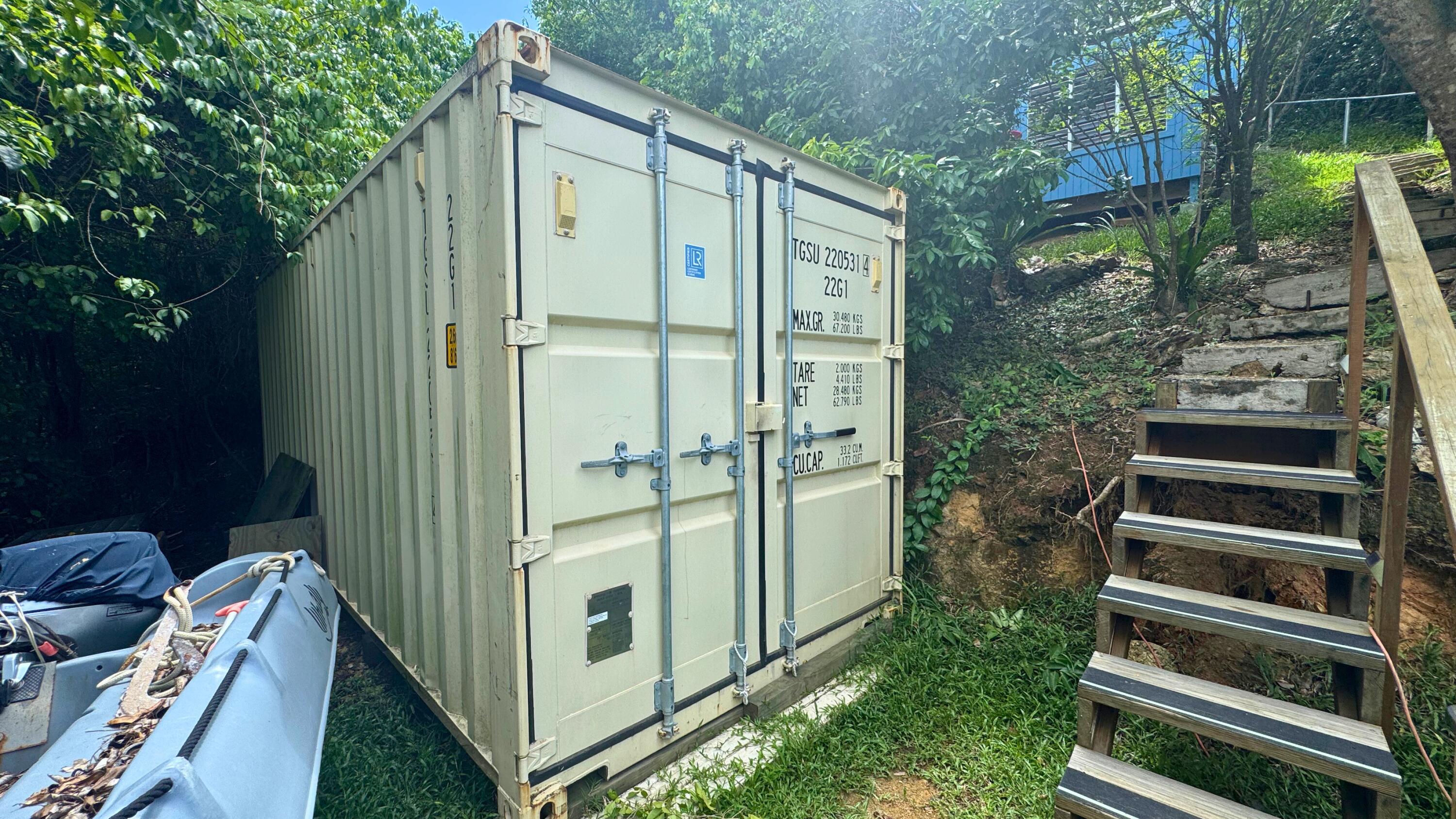 White shipping container on a wooded hillside beside wooden stairs, with kayaks nearby.