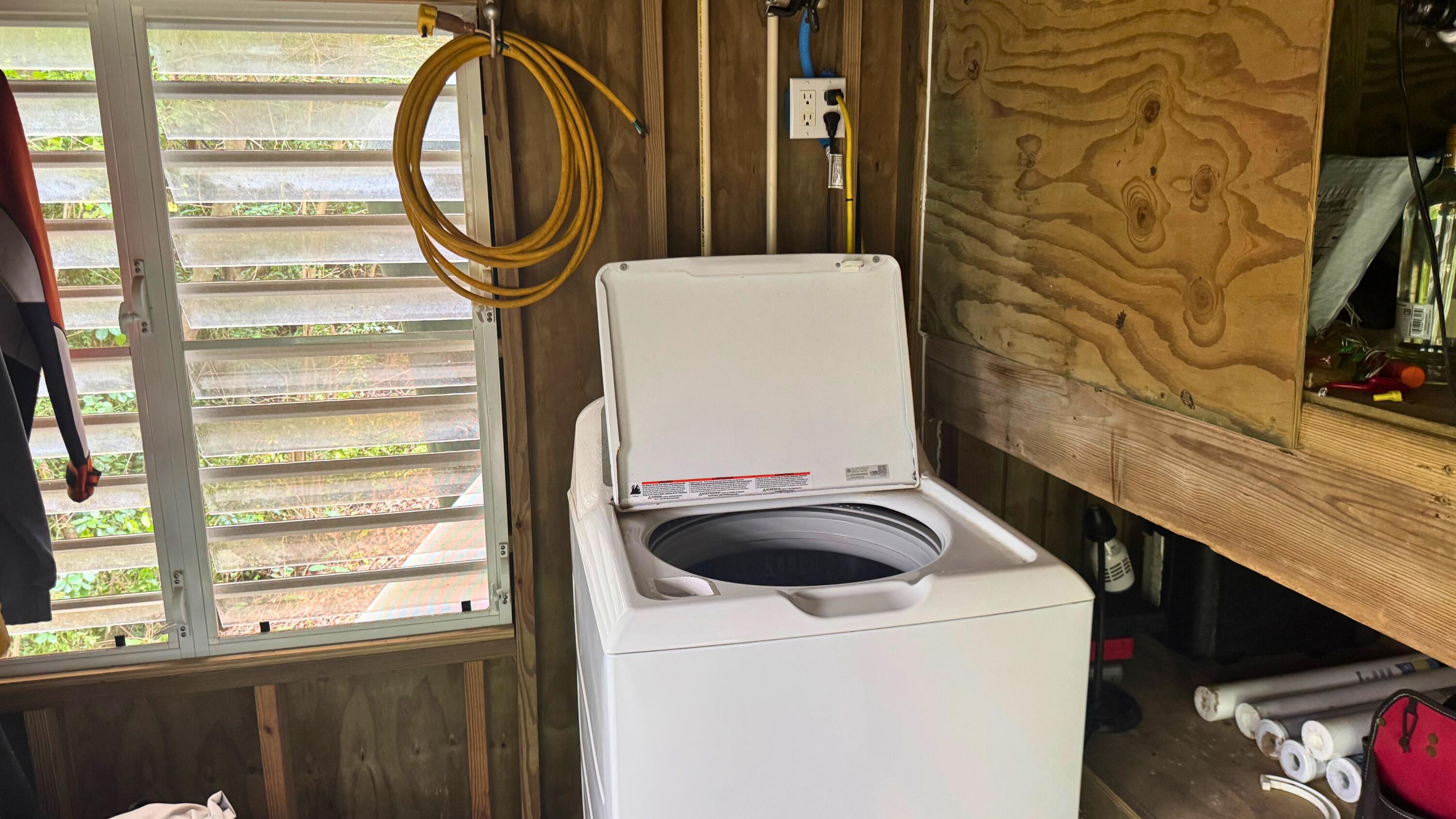 Open top-loading washer in a rustic utility room beside a louvered window.