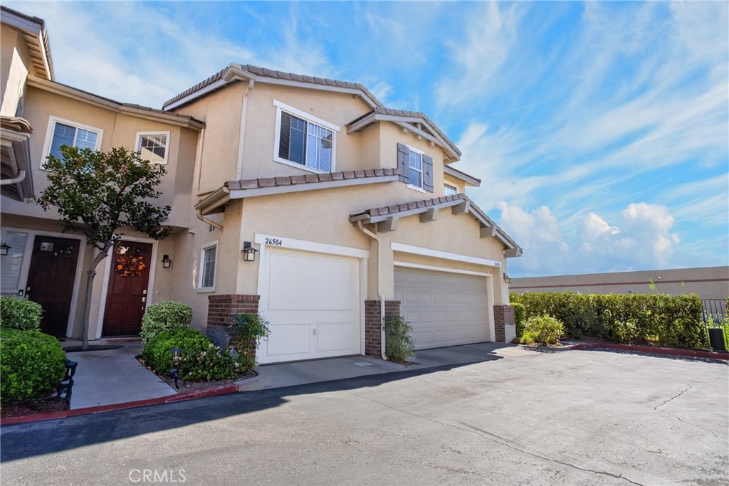 Two-story beige townhouse with attached garages, a landscaped entry, and a bright blue sky.