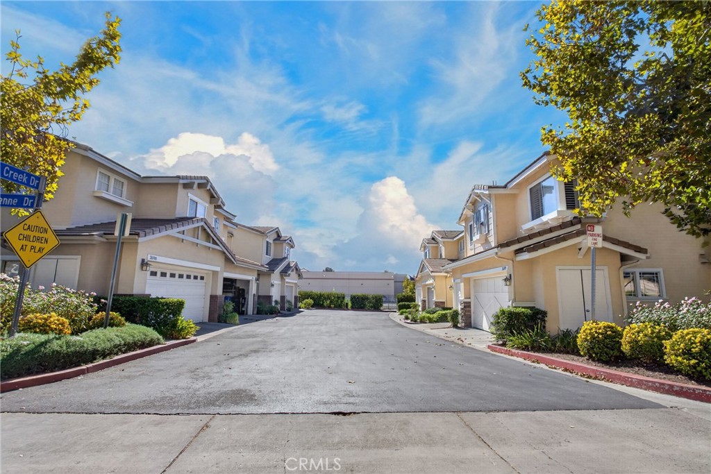 Residential complex street with attached homes, garages, landscaped yards, and a clear blue sky.