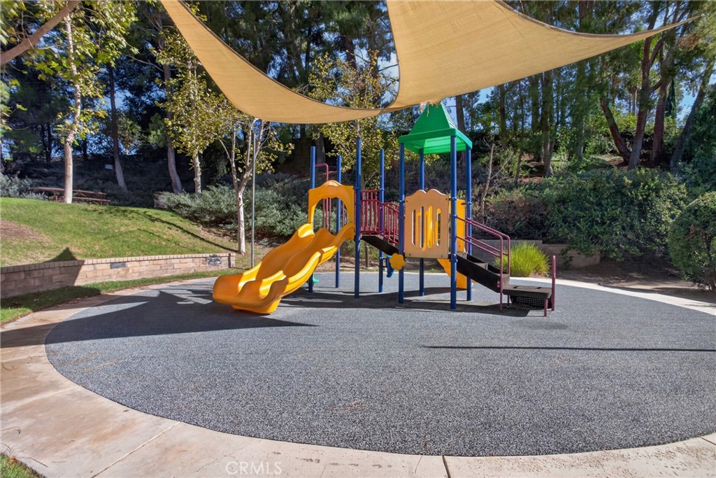 Playground with colorful slides beneath a tan shade canopy, surrounded by trees.