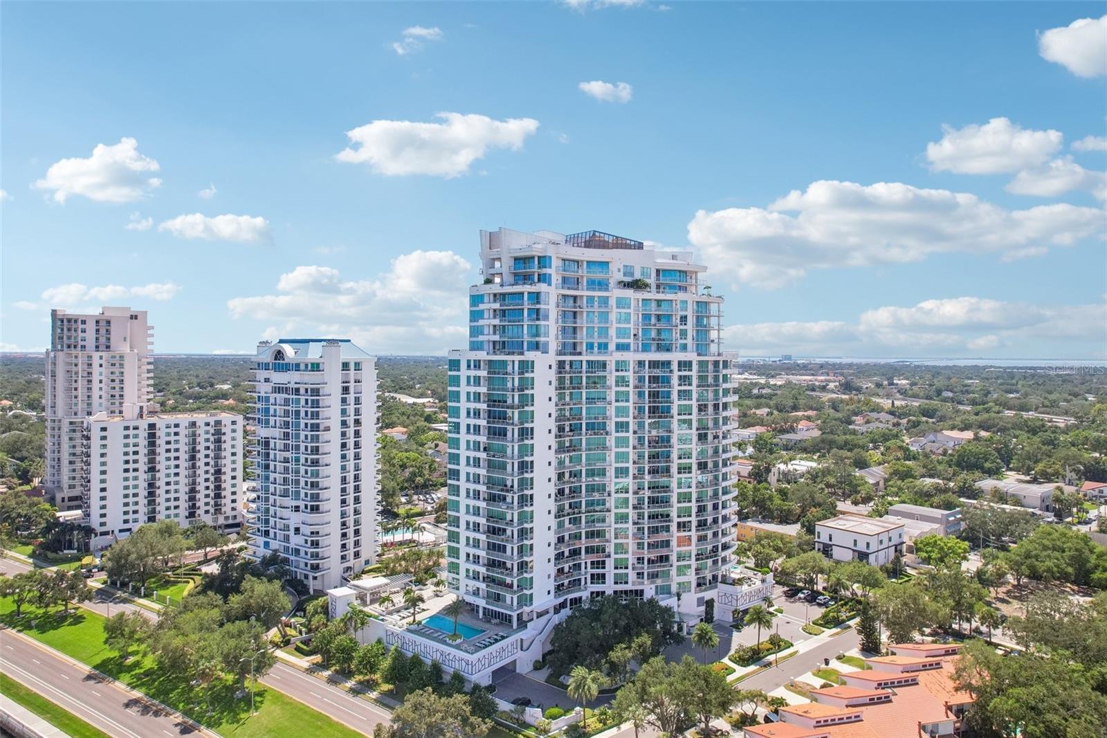 Aerial view of white condominium towers with glass balconies amid tree-lined streets and homes.