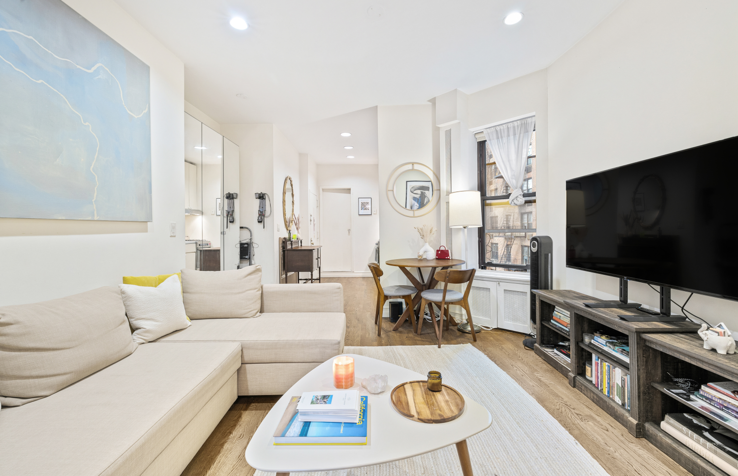 Open-plan living room with a cream sectional, wood floors, and dining table beside tall windows.