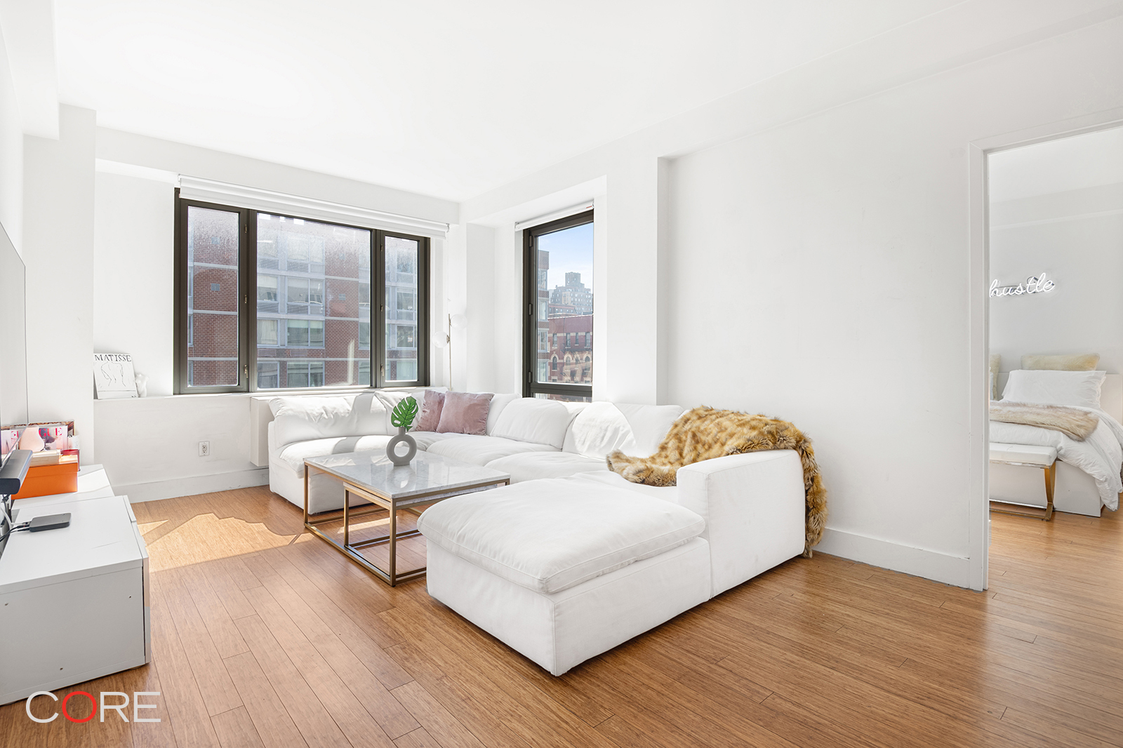 Bright apartment living room with a white sectional, large windows, and a view into the bedroom.