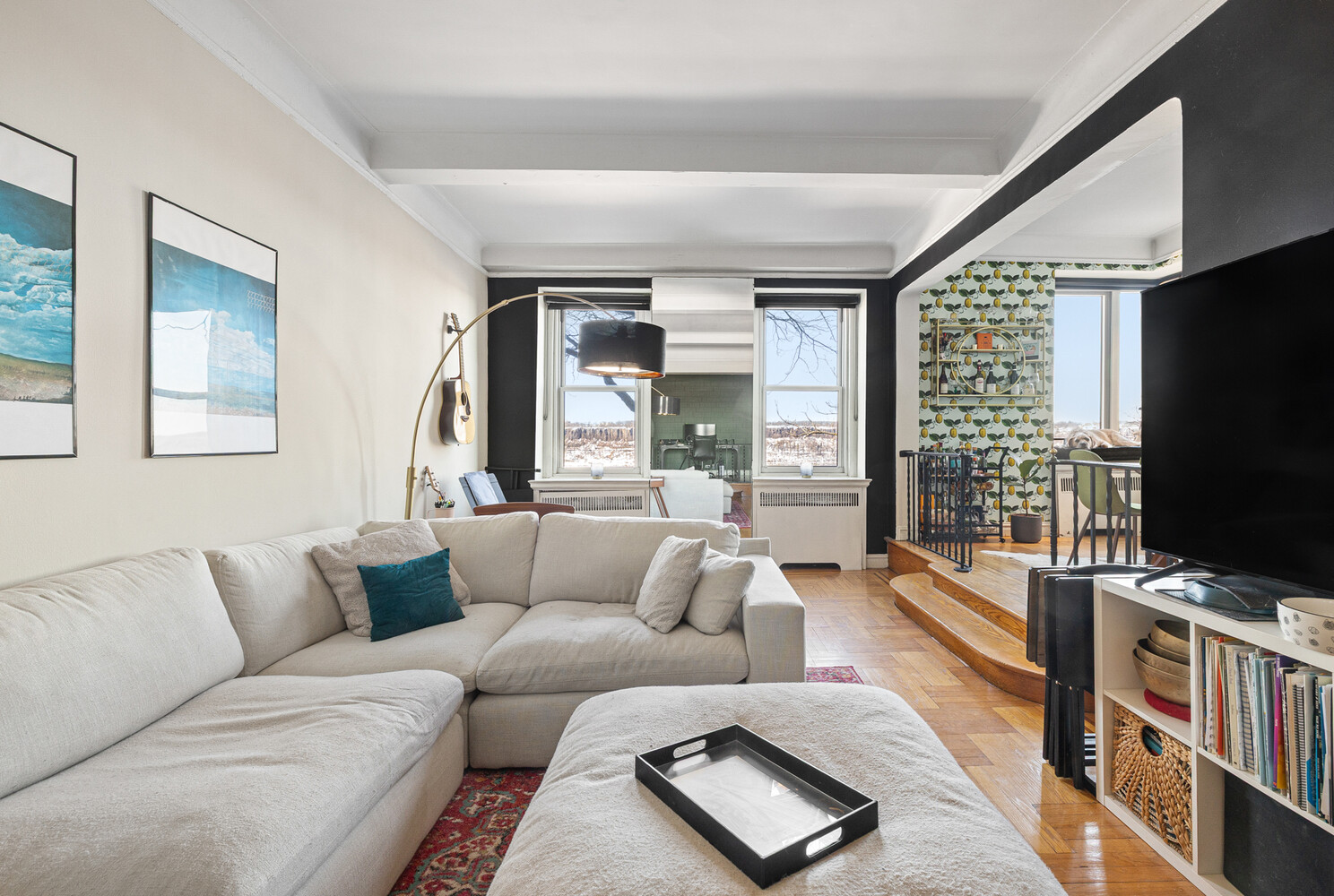 Living room with a white sectional, parquet floors, large windows, and a raised dining nook with citrus-patterned wallpaper.