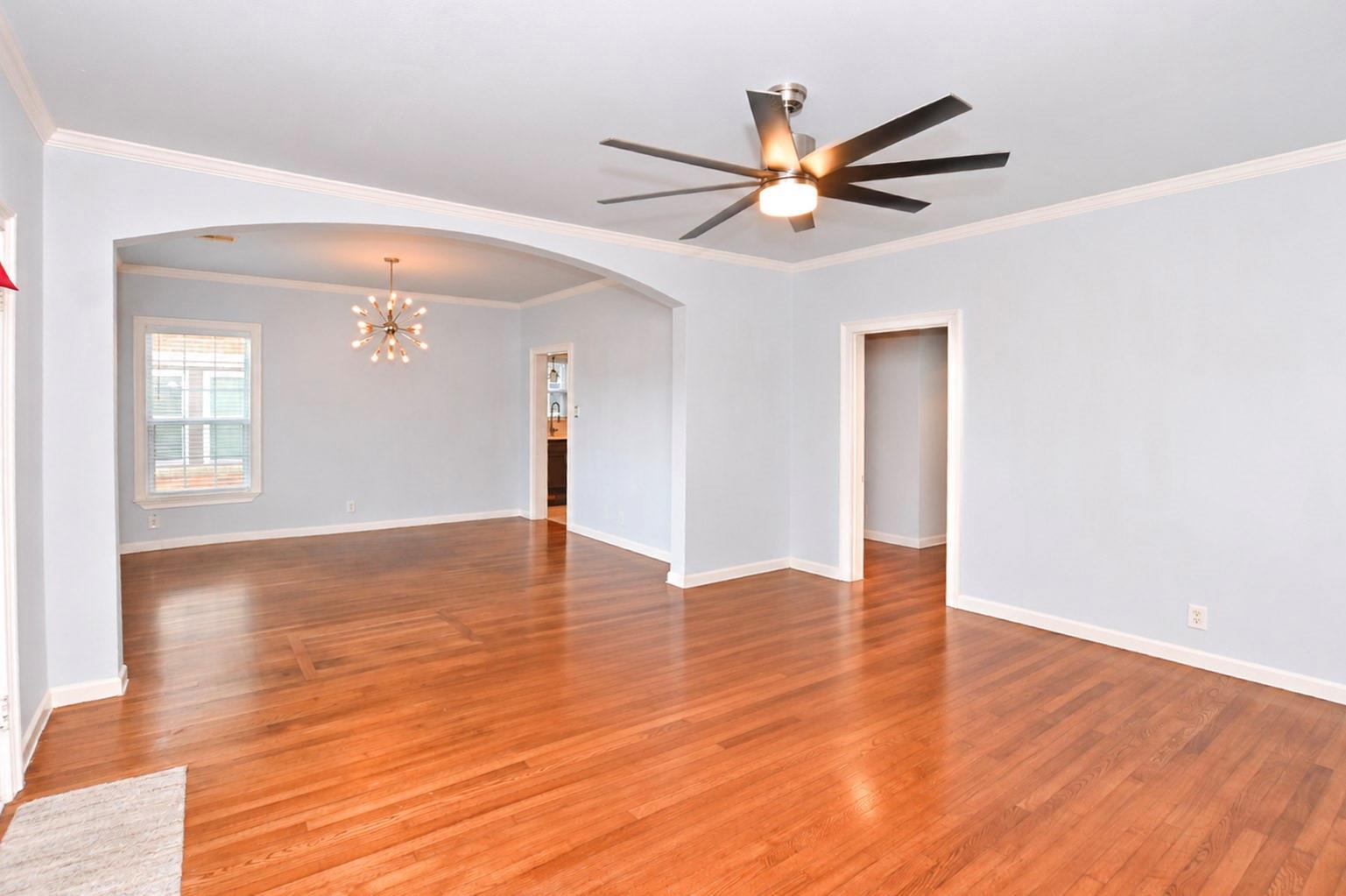 Empty living room with hardwood floors, a large ceiling fan, and an arched opening to a room with a chandelier.