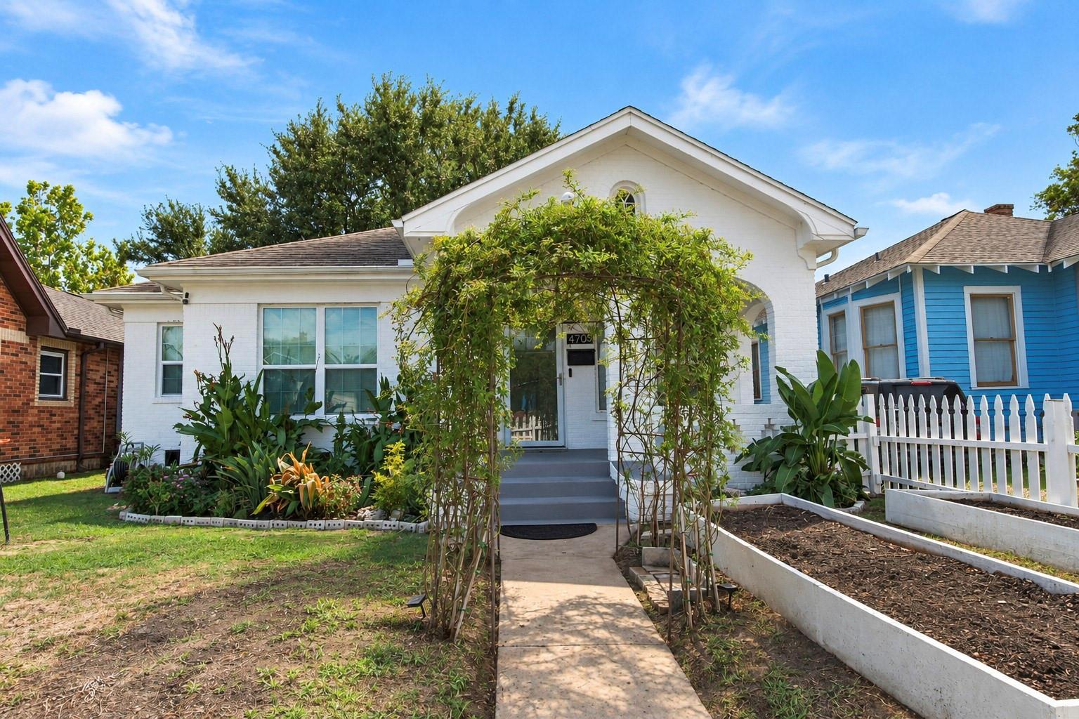 White brick house with a vine-covered entry arbor, raised garden beds, and a white picket fence.