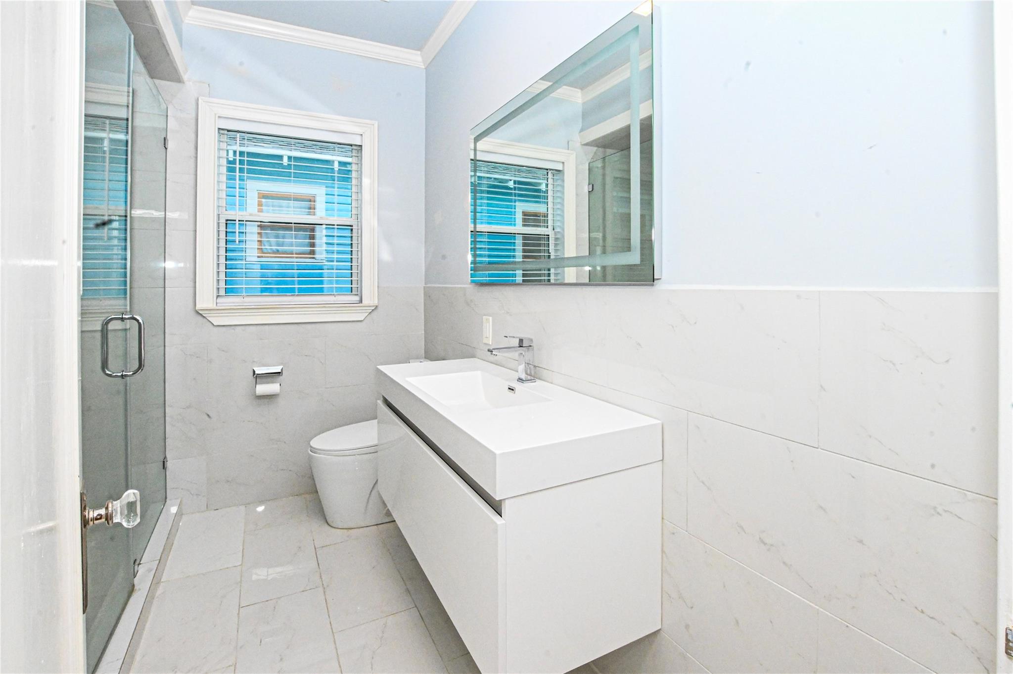 Modern bathroom with marble-look tile, glass shower, floating vanity, and window with blinds.