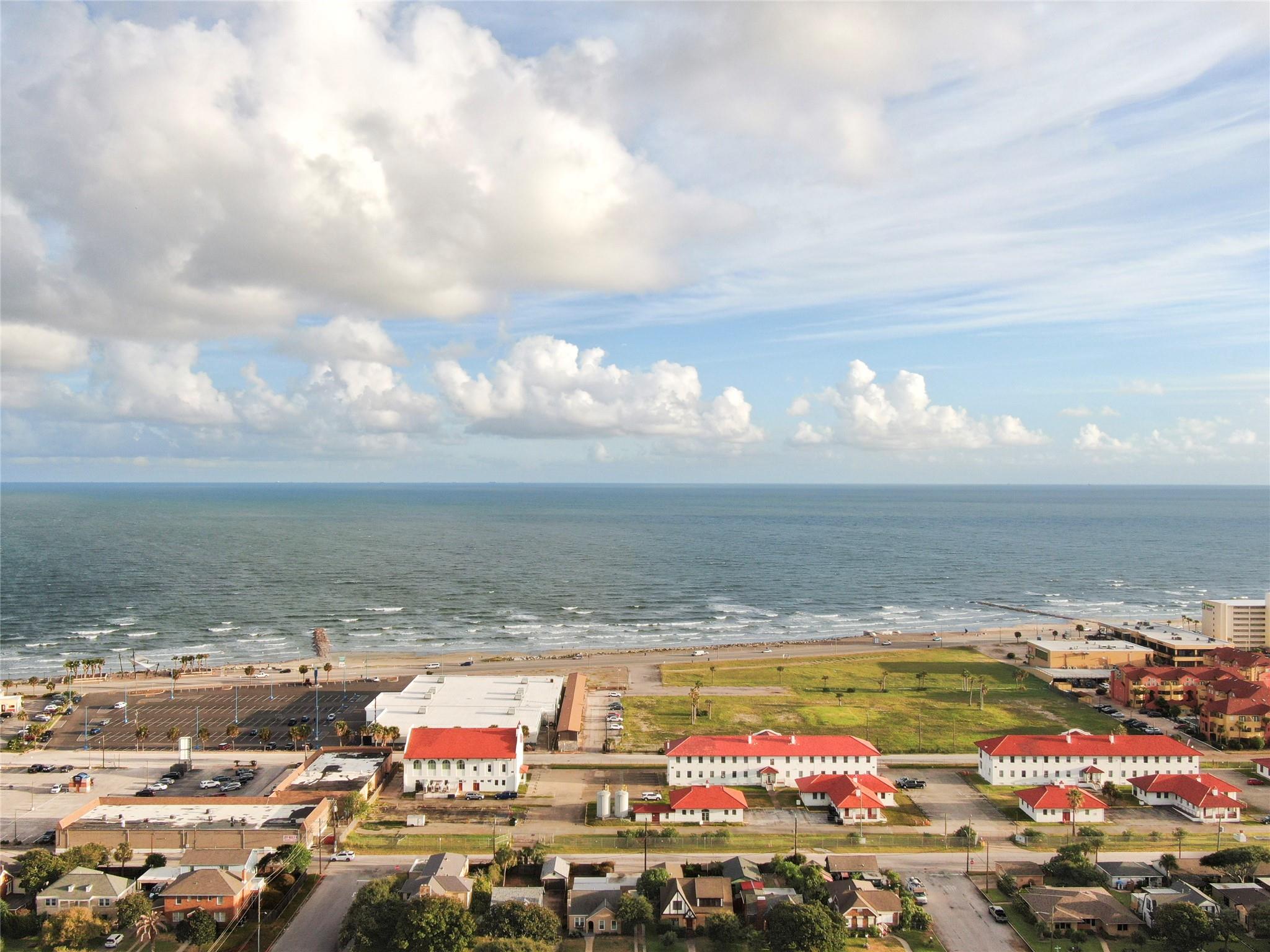 Aerial view of the Gulf shoreline, beach, and nearby red-roofed buildings beneath a partly cloudy sky.