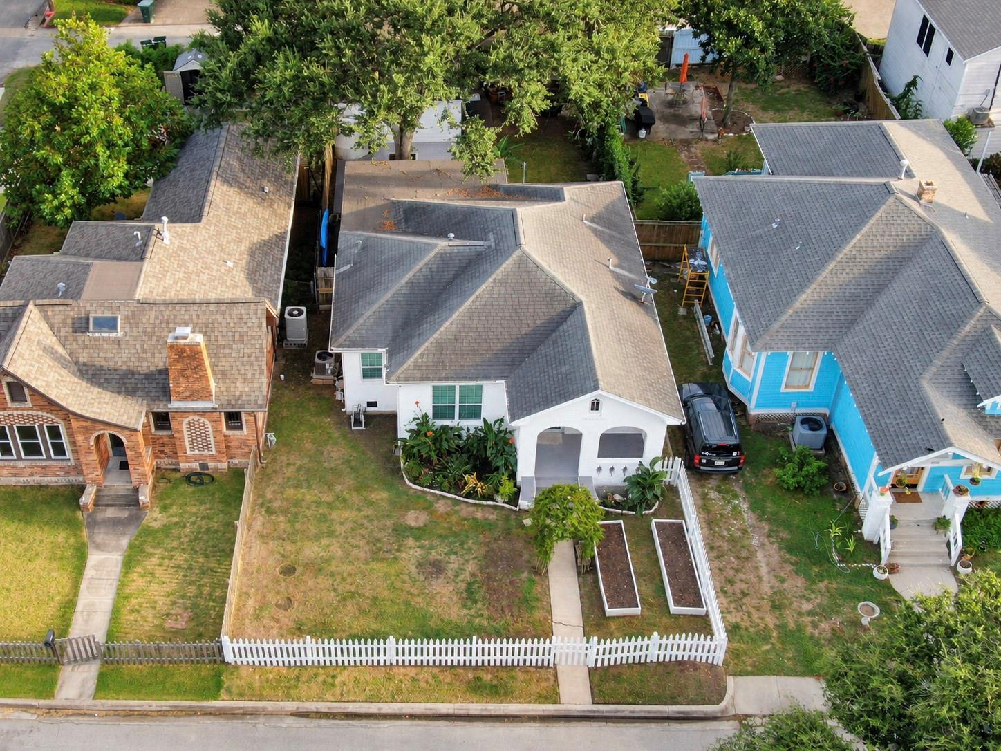 Aerial view of a white house with a gray roof, fenced front yard, and raised garden beds.
