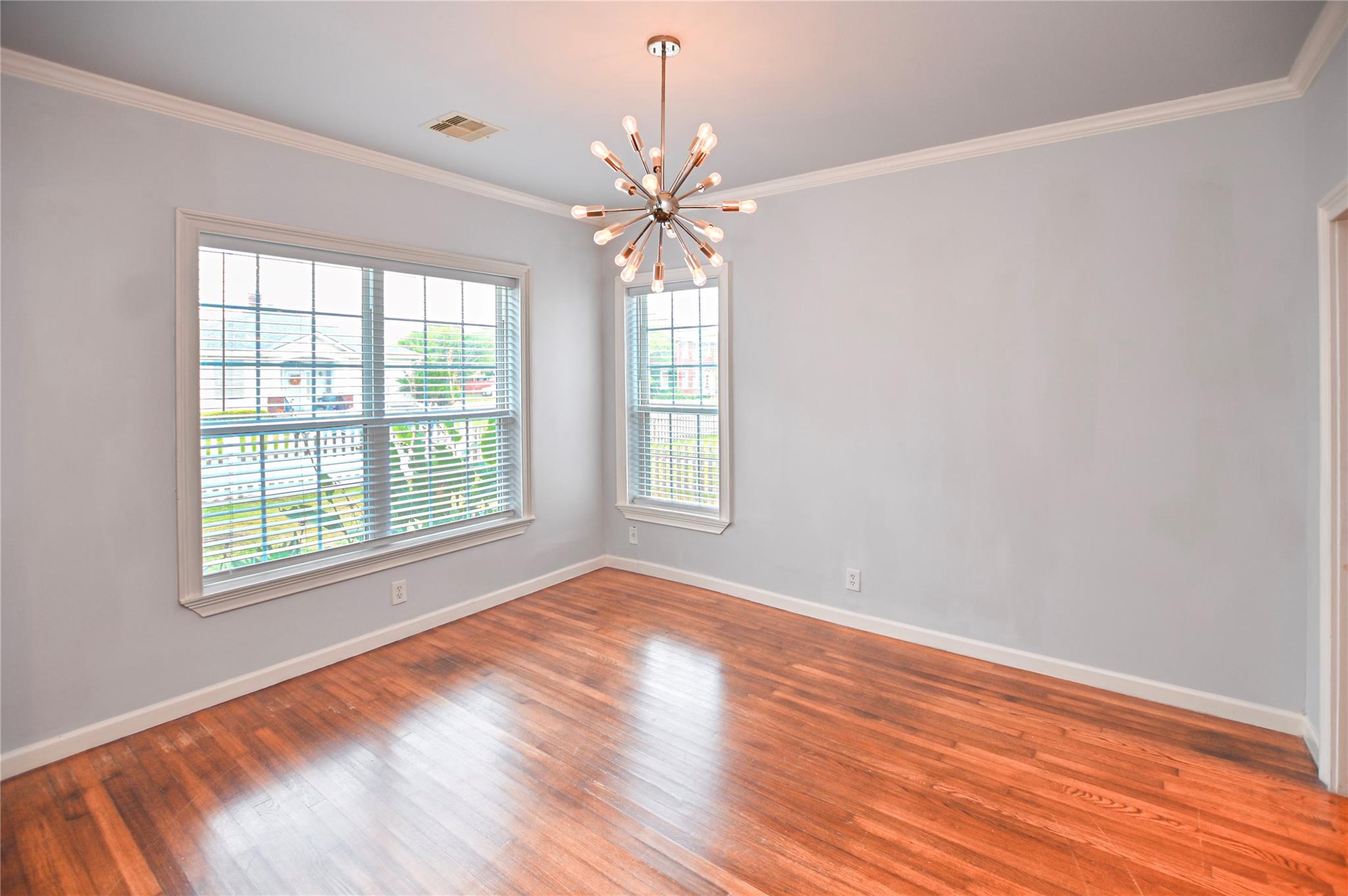Empty room with warm hardwood floors, two windows with blinds, and a starburst chandelier.