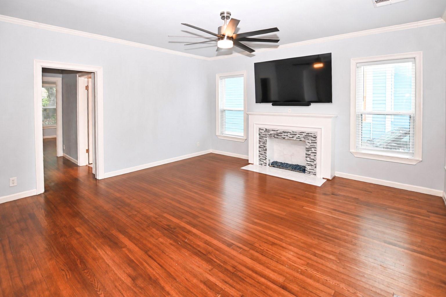 Vacant living room with hardwood floors, a tiled fireplace, wall-mounted TV, and ceiling fan.