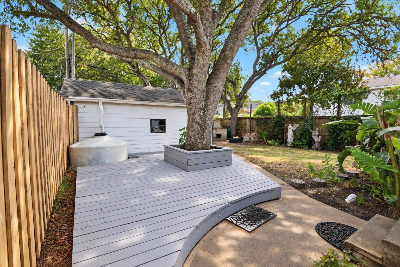 Backyard deck wraps around a mature tree, with a white outbuilding and fenced garden.