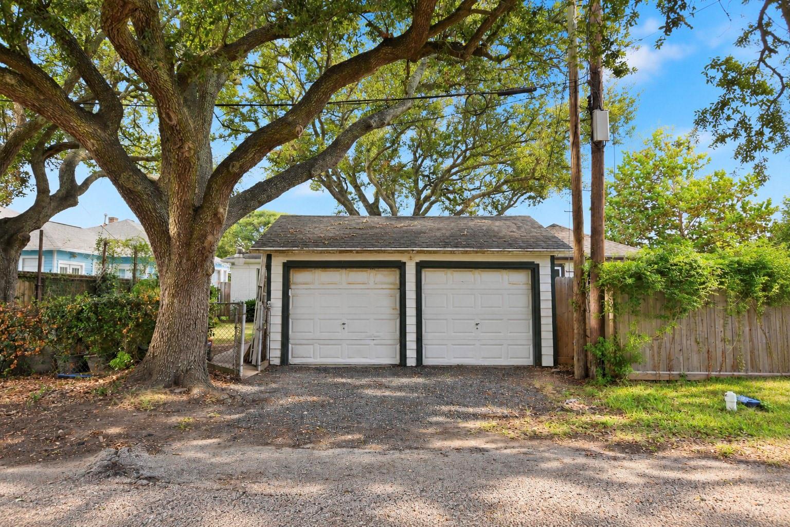 Detached white two-car garage beneath a large leafy tree, viewed from the gravel driveway.