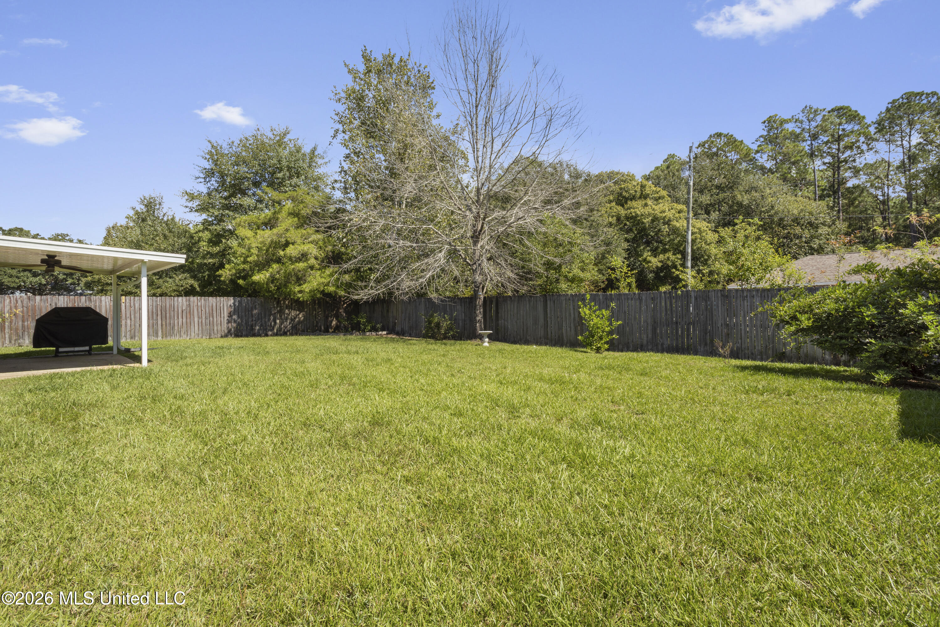 Fenced grassy backyard with a covered patio and grill beneath mature trees.