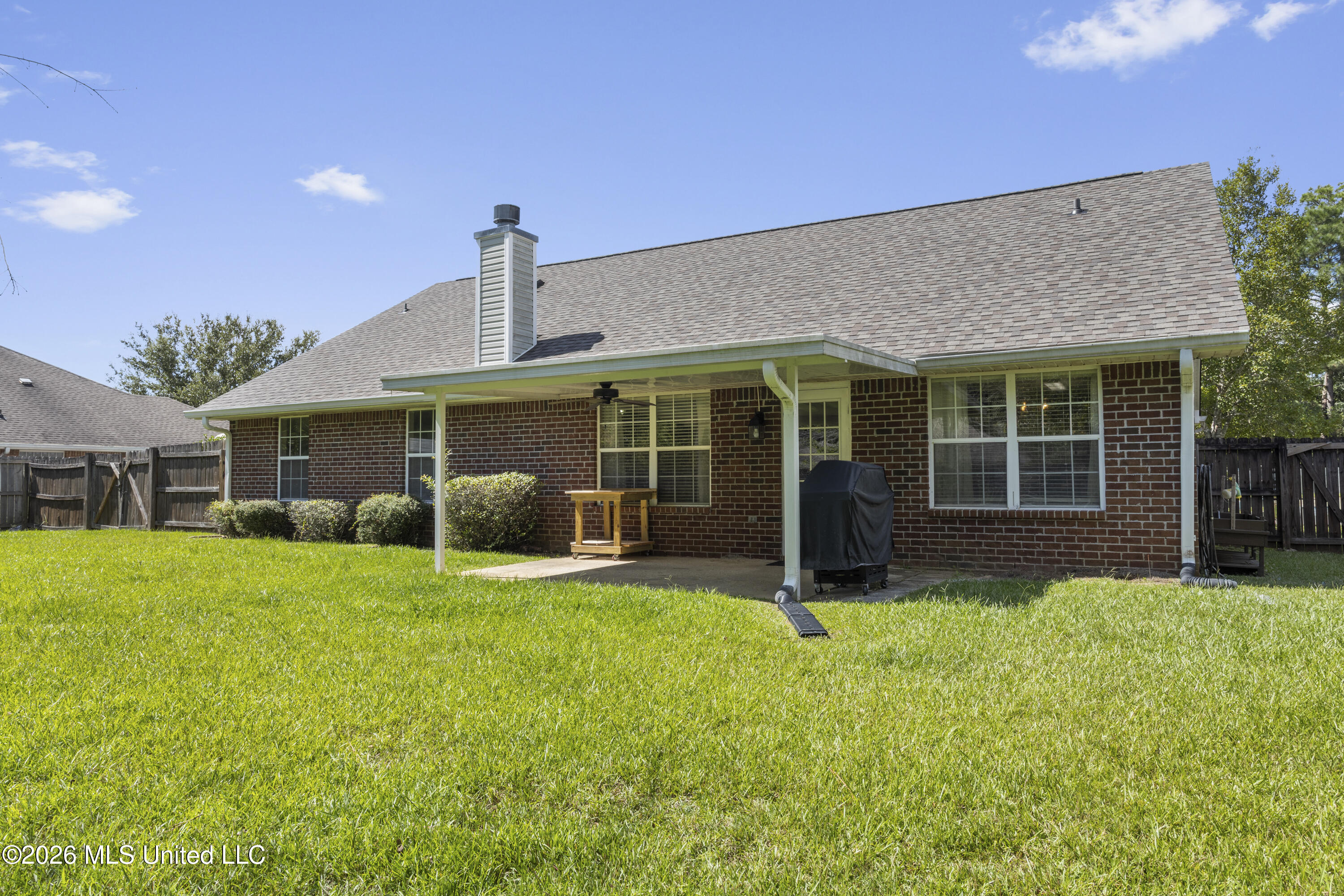 Brick home’s rear exterior with a covered patio, grill, fenced yard, and green lawn.