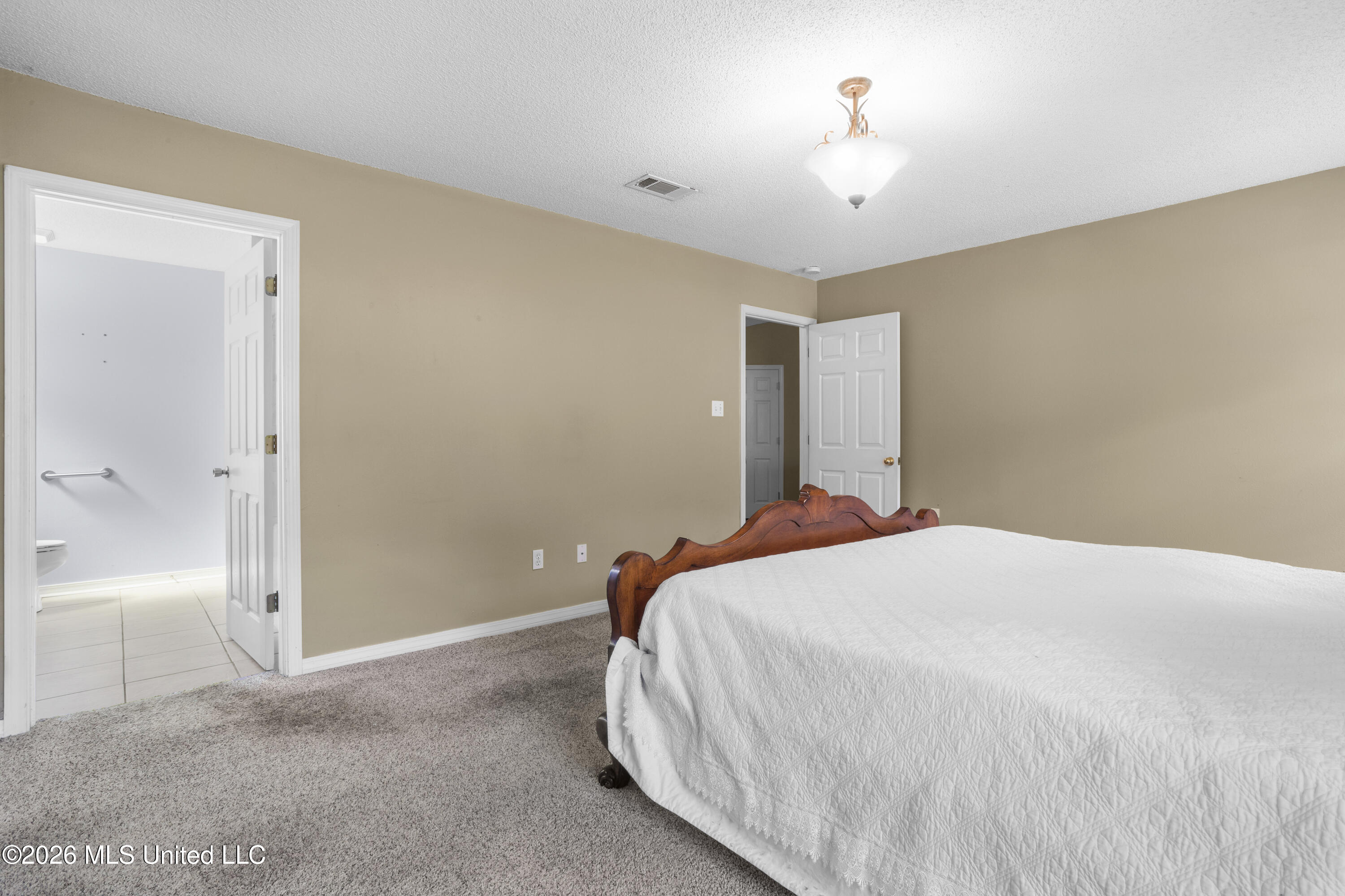 Bedroom with a wooden bed and white quilt, gray carpet, and an open doorway to the bathroom.