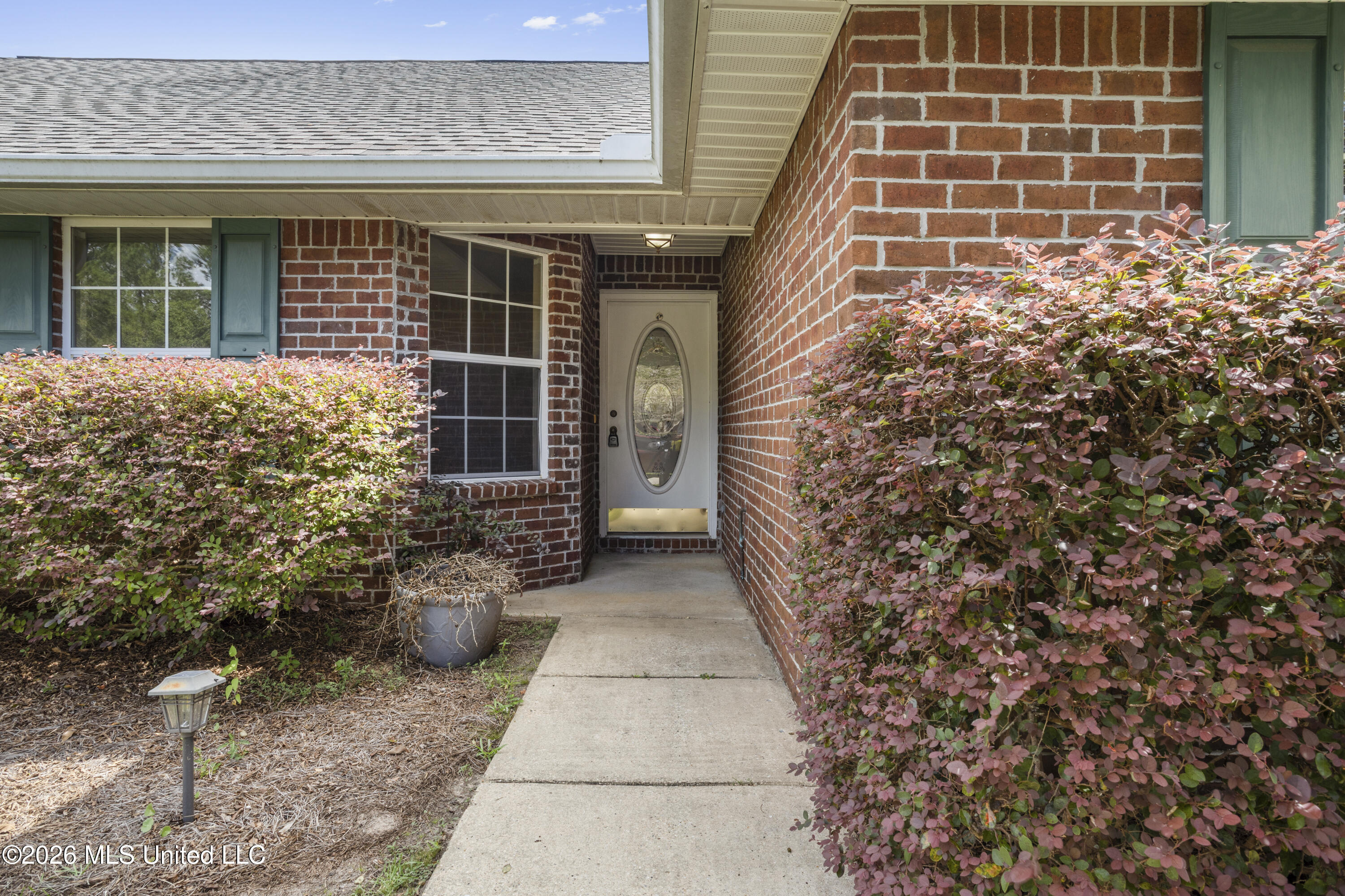 Brick home’s covered front entrance with an oval-glass door and pink-leaved shrubs along the walkway.
