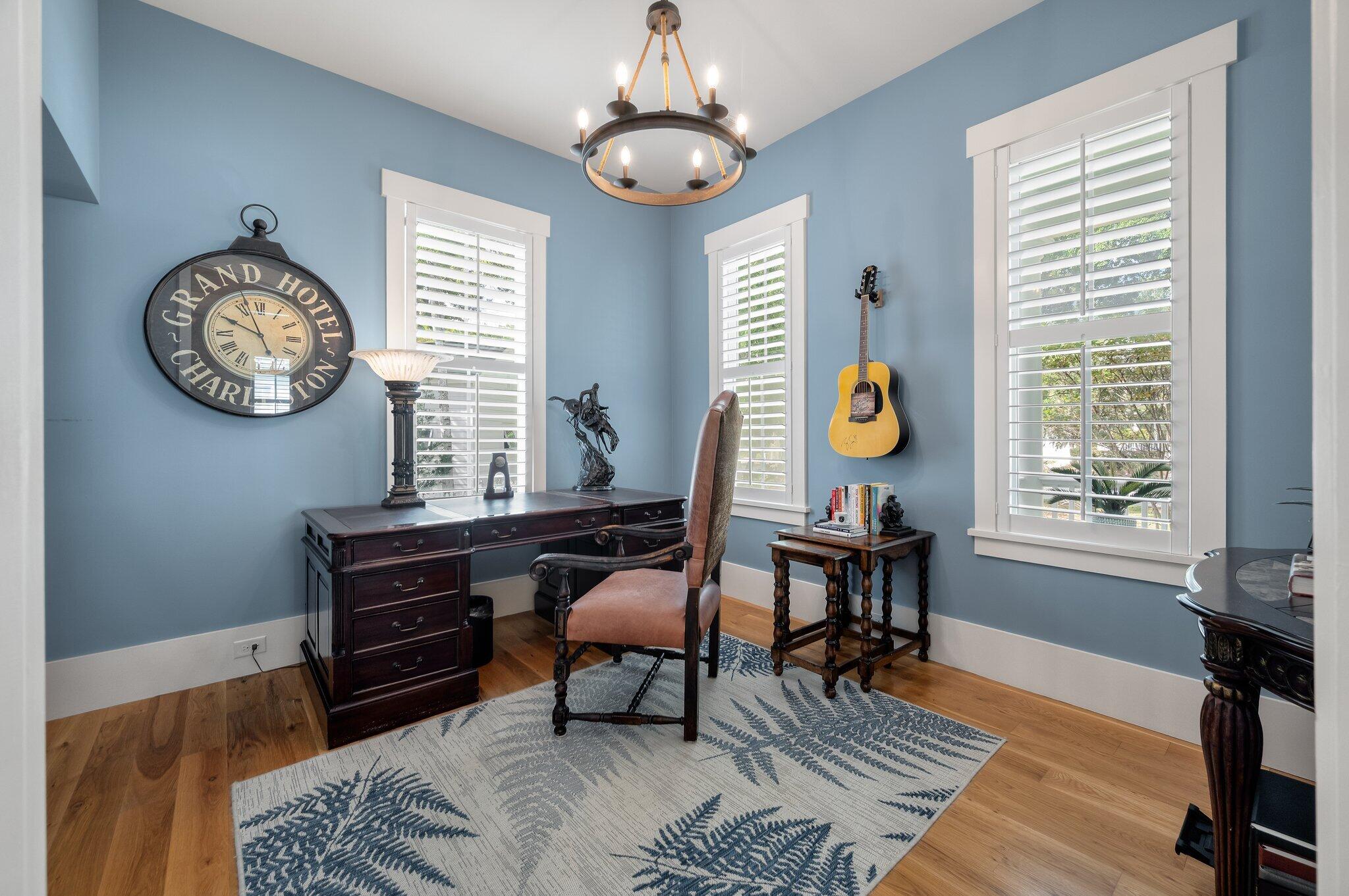 Blue home office with plantation shutters, a dark wood desk, leather chair, and wall-mounted guitar.