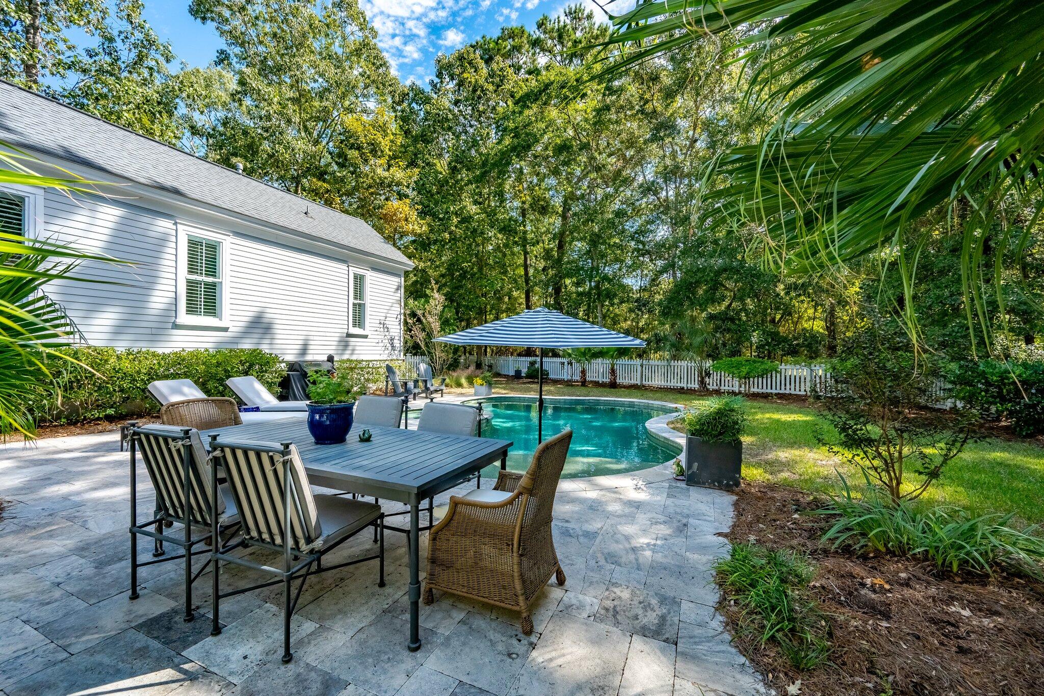 Backyard pool beside a stone patio with dining furniture and a striped umbrella.