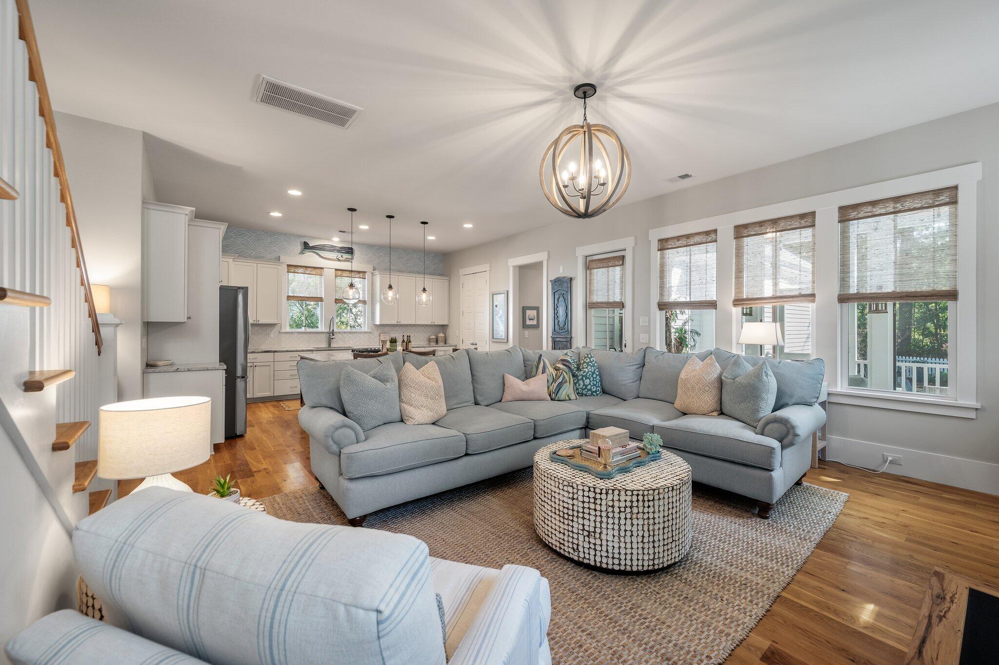 Open-plan living room with a gray sectional, patterned coffee table, tall shaded windows, and adjoining kitchen.