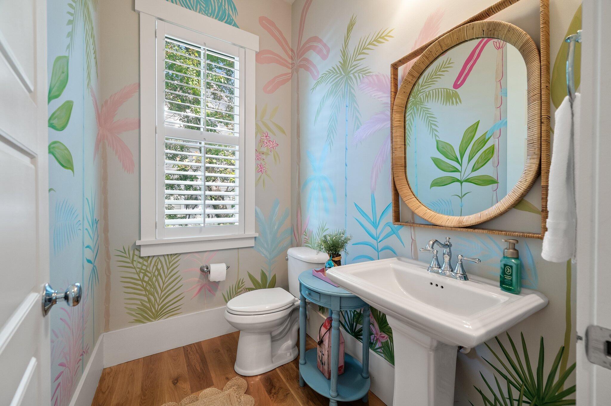Bathroom with colorful tropical mural wallpaper, a rattan-framed mirror, and white pedestal sink.
