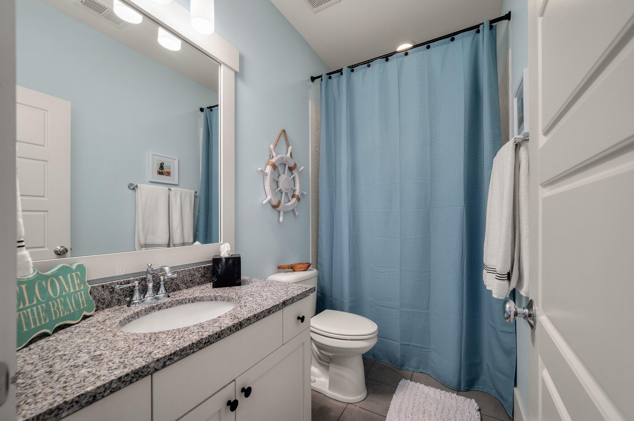 Bathroom with a granite vanity and blue shower curtain, decorated with a nautical ship's wheel.