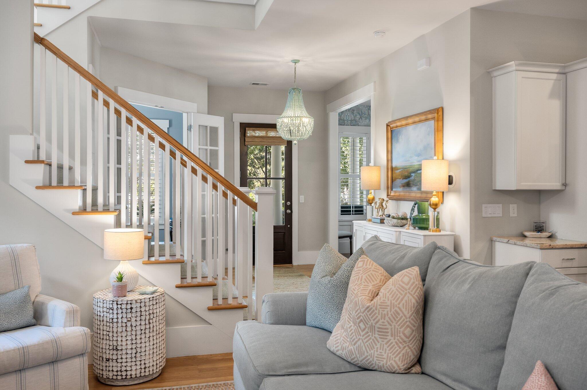 Living room with a gray sofa, white staircase with wood accents, and a chandelier near the entry.