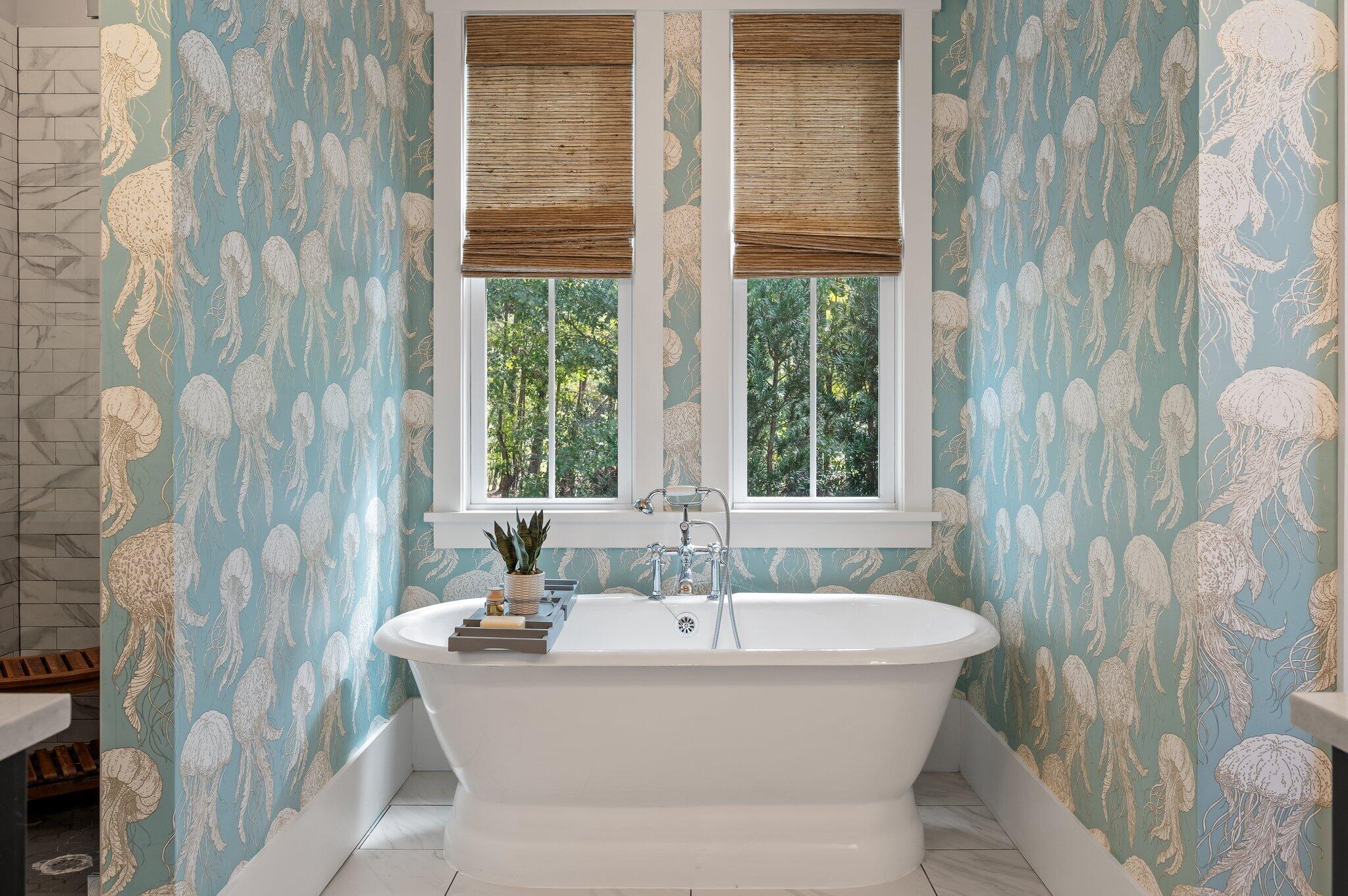 Freestanding soaking tub against blue jellyfish wallpaper, beneath two shaded windows.