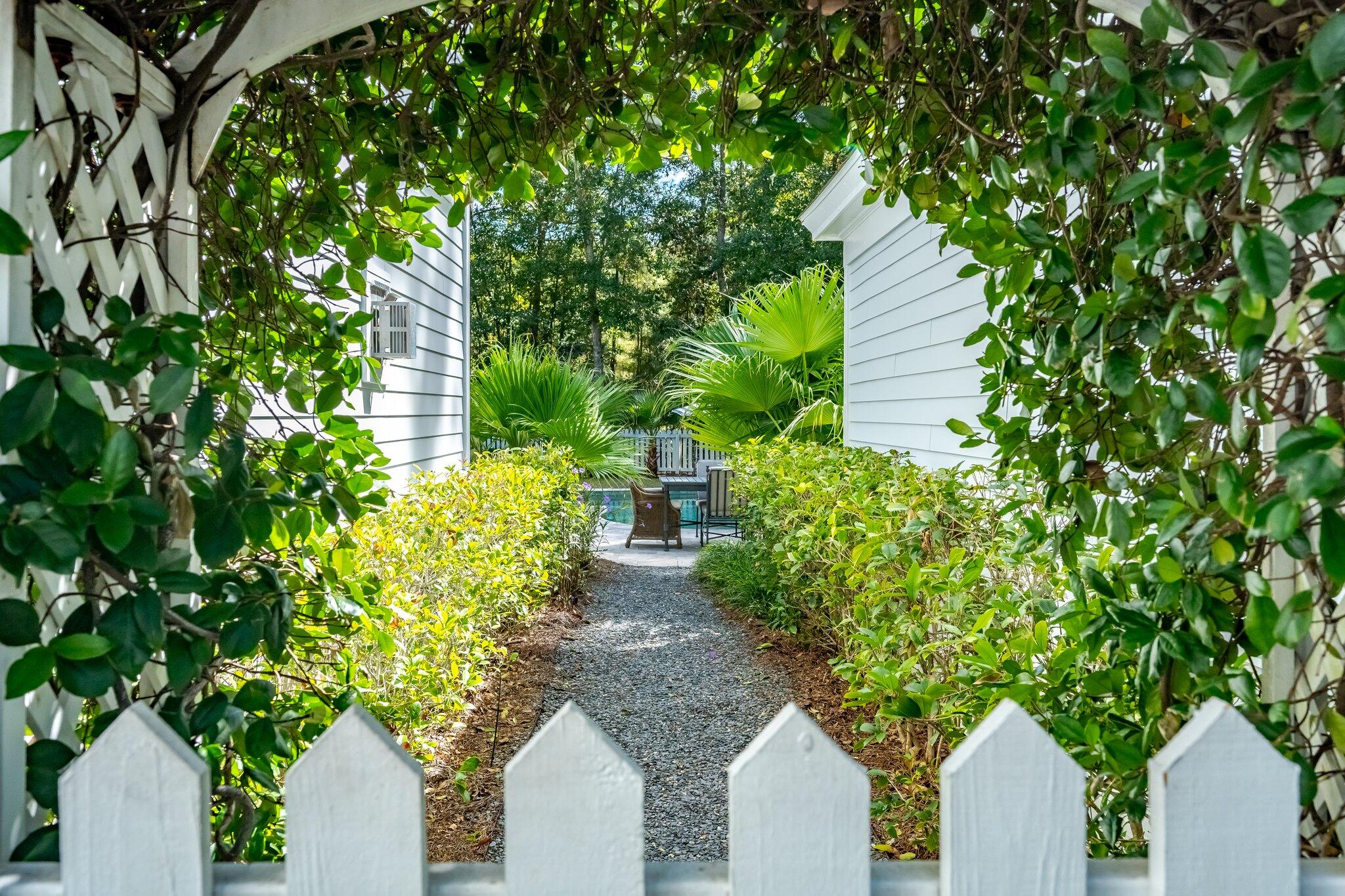 Gravel path through lush palms and shrubs leads to a patio with chairs between white-sided walls.