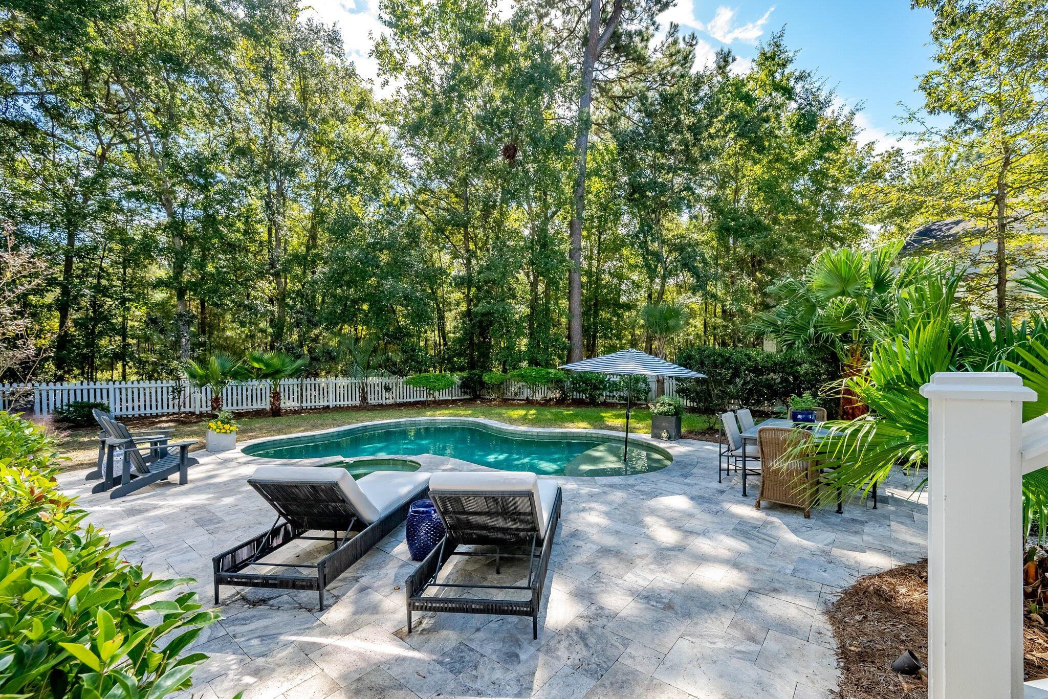 Backyard pool with a stone patio, lounge chairs, striped umbrella, and wooded surroundings.