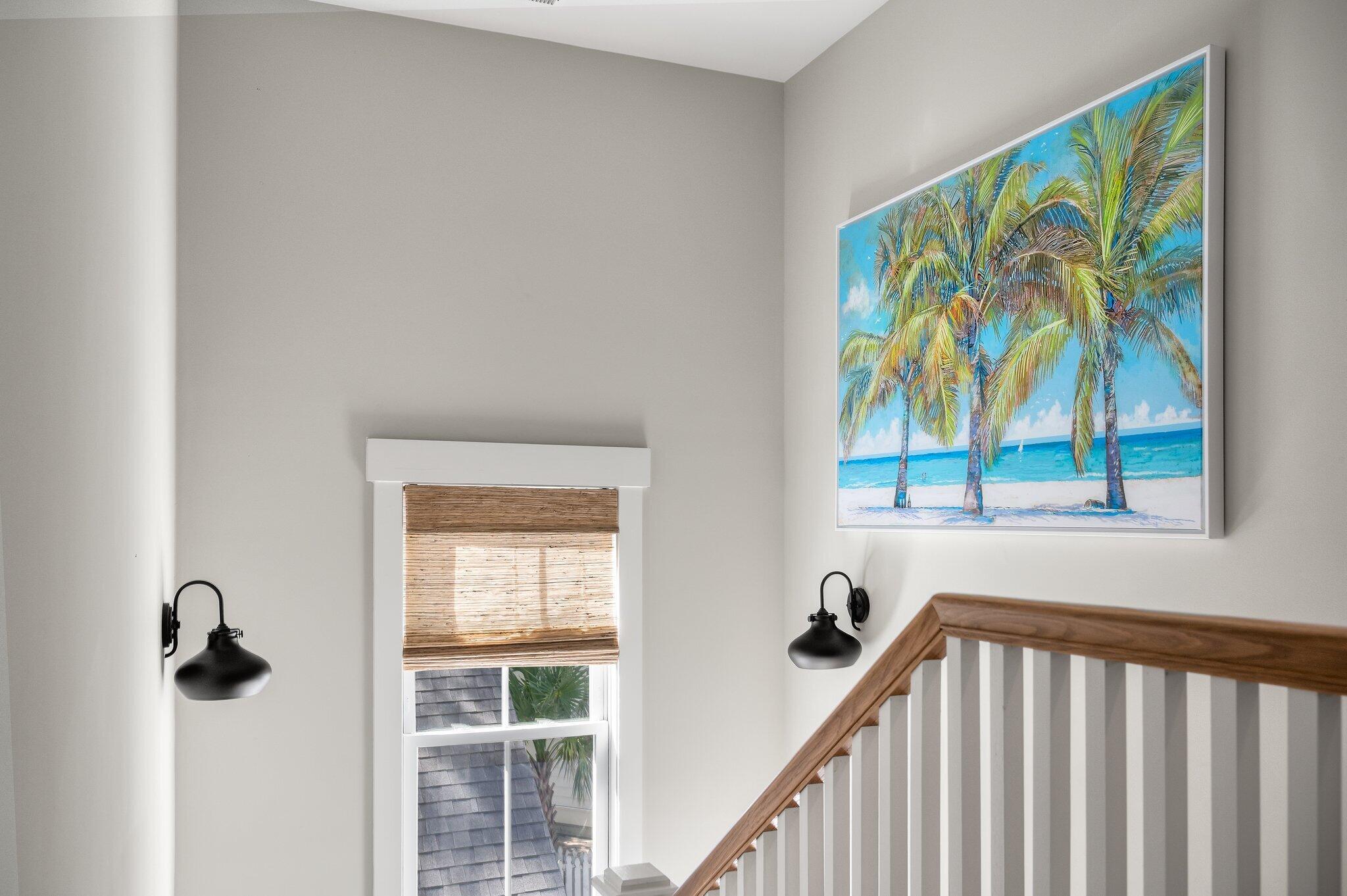 Interior stairwell with a wood handrail, woven window shade, and framed tropical palm-tree artwork.
