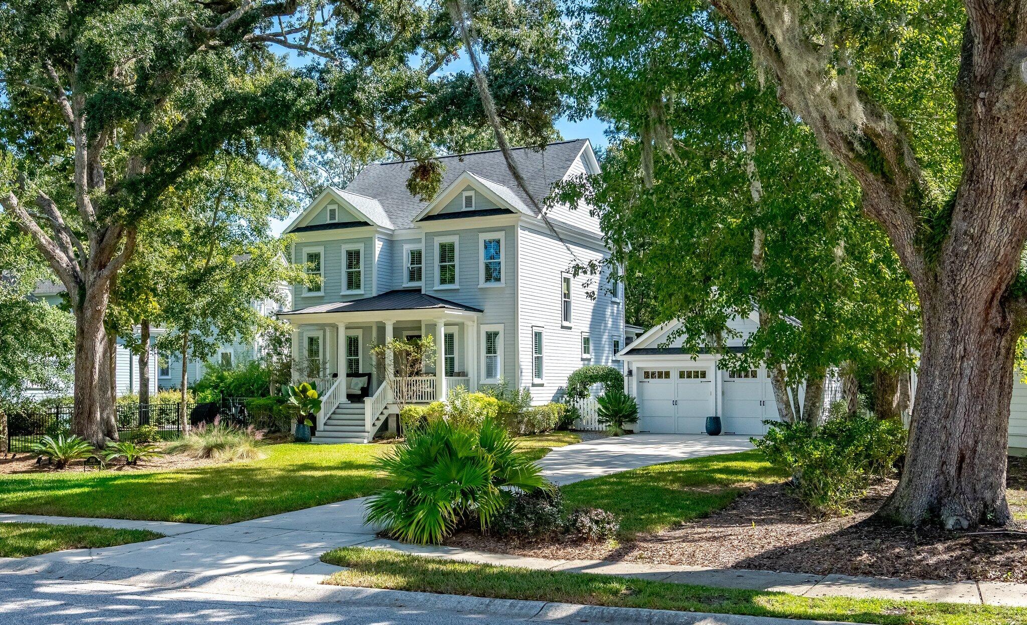 Two-story light-colored house with a covered porch and double garage beneath mature moss-draped trees.