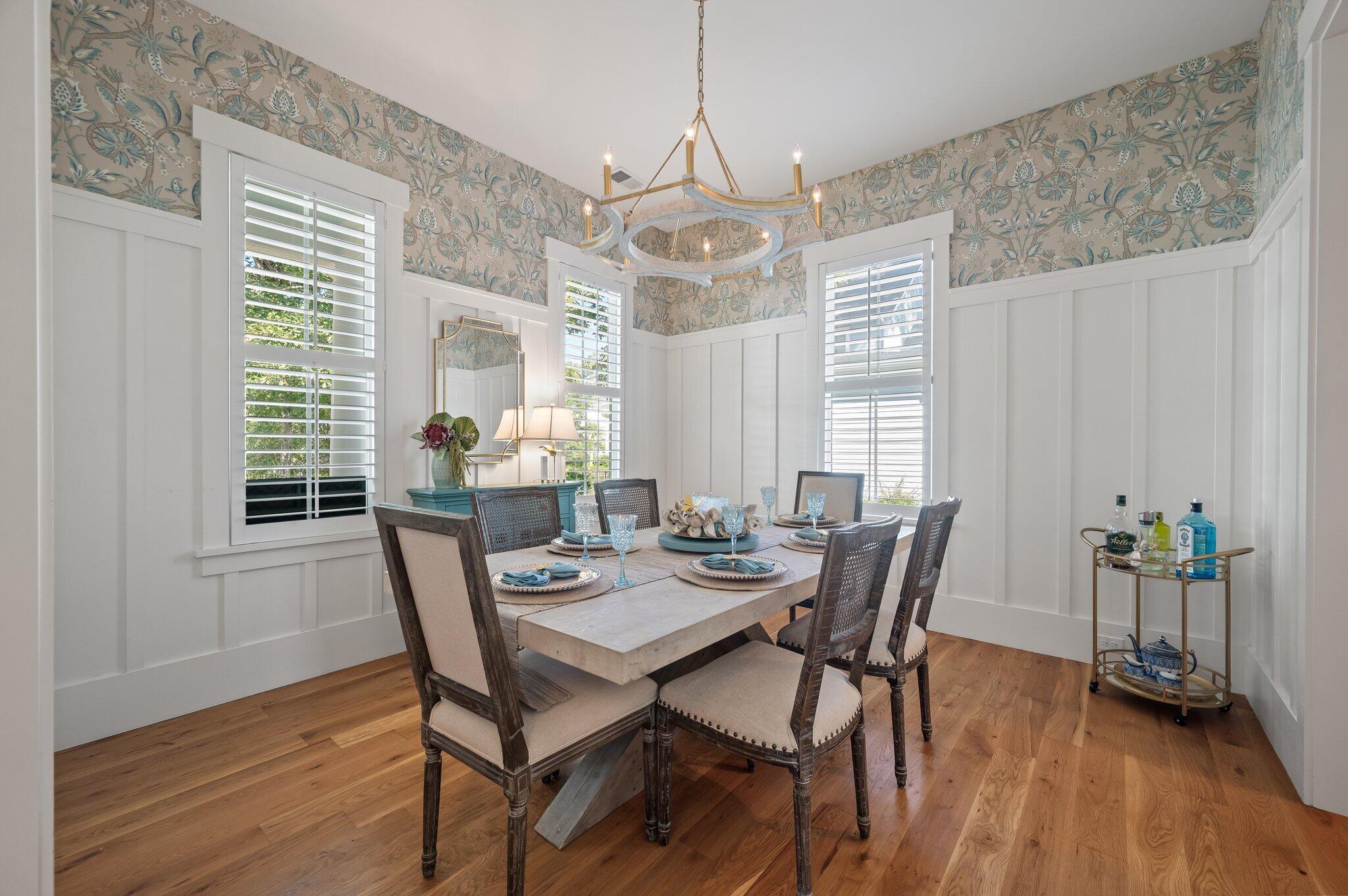 Dining room with a set table, floral wallpaper, white paneling, and shuttered windows.