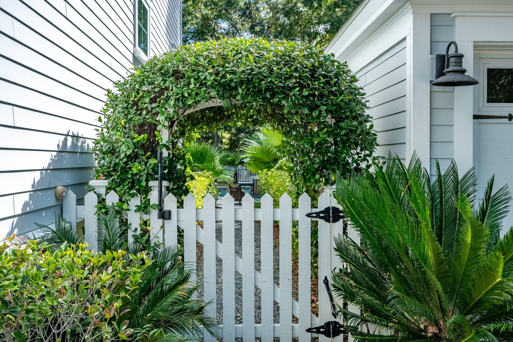 White picket gate beneath a leafy arbor beside a light-colored house, surrounded by tropical plants.