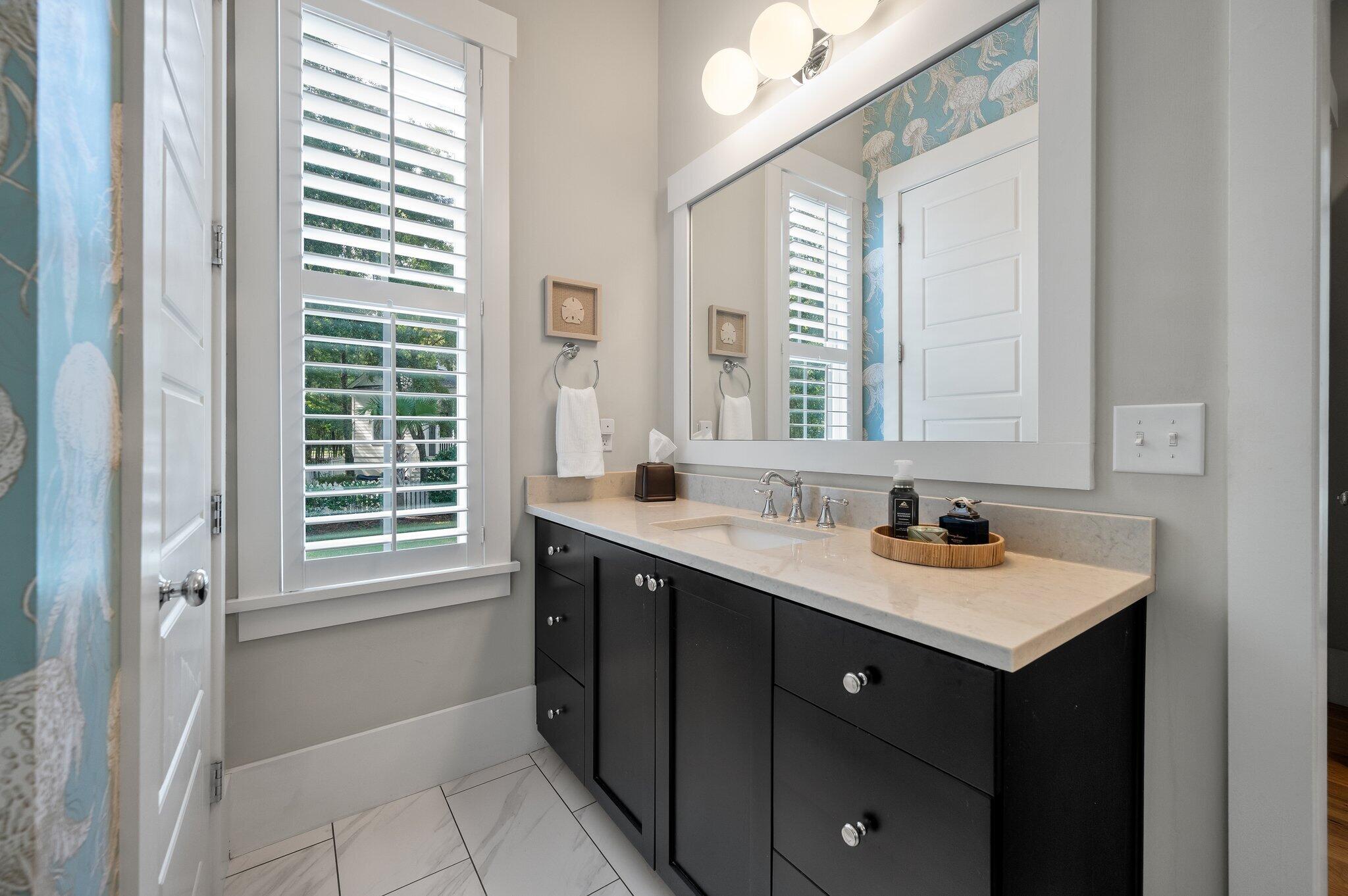 Bathroom vanity with dark cabinets, white stone counter, large mirror, and plantation-shuttered window.