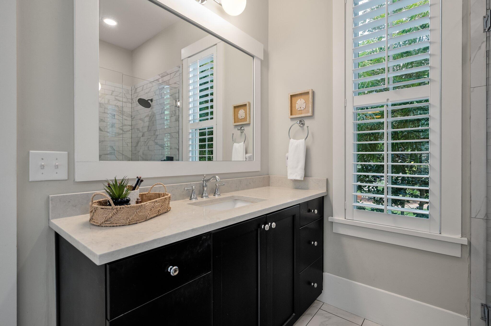 Bathroom vanity with dark cabinets, a large mirror, plantation shutters, and a tiled walk-in shower.