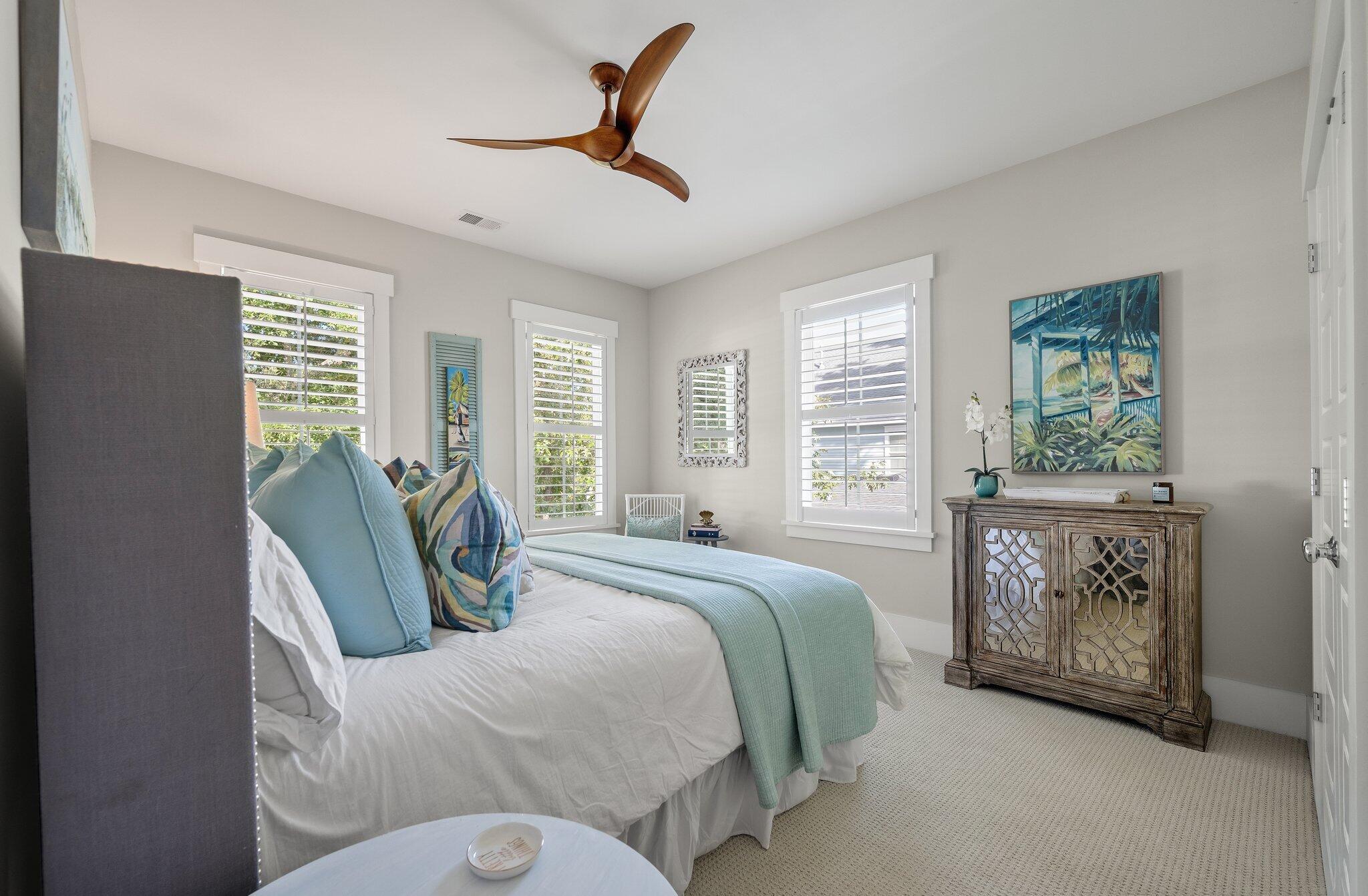Staged bedroom with pale aqua bedding, shuttered windows, and a carved wooden cabinet beneath tropical artwork.