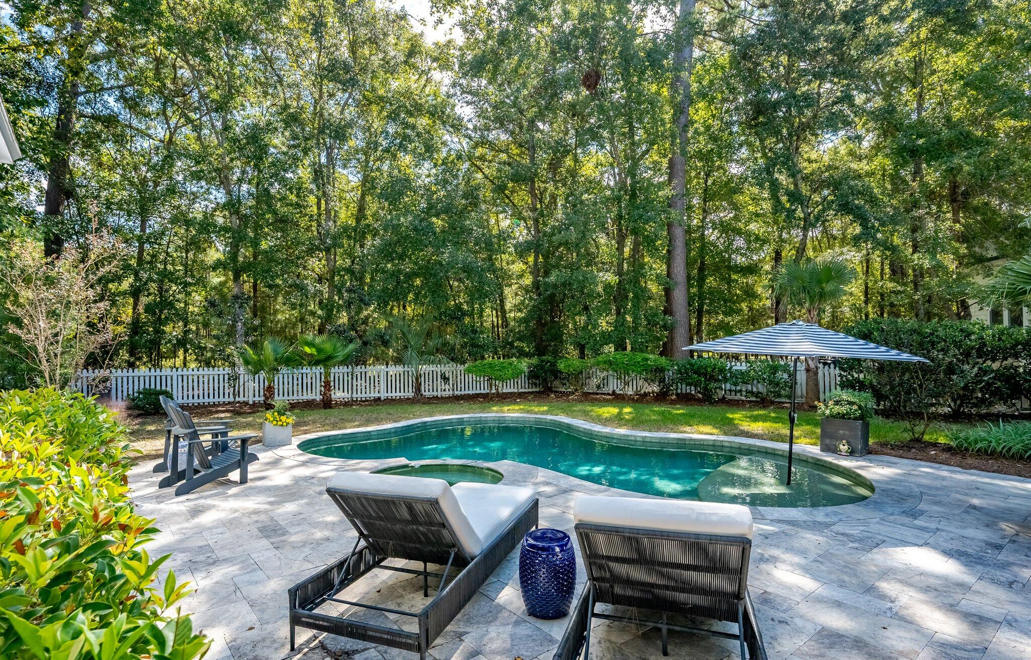 Curved backyard pool bordered by a stone patio, lounge chairs, striped umbrella, and wooded privacy fence.