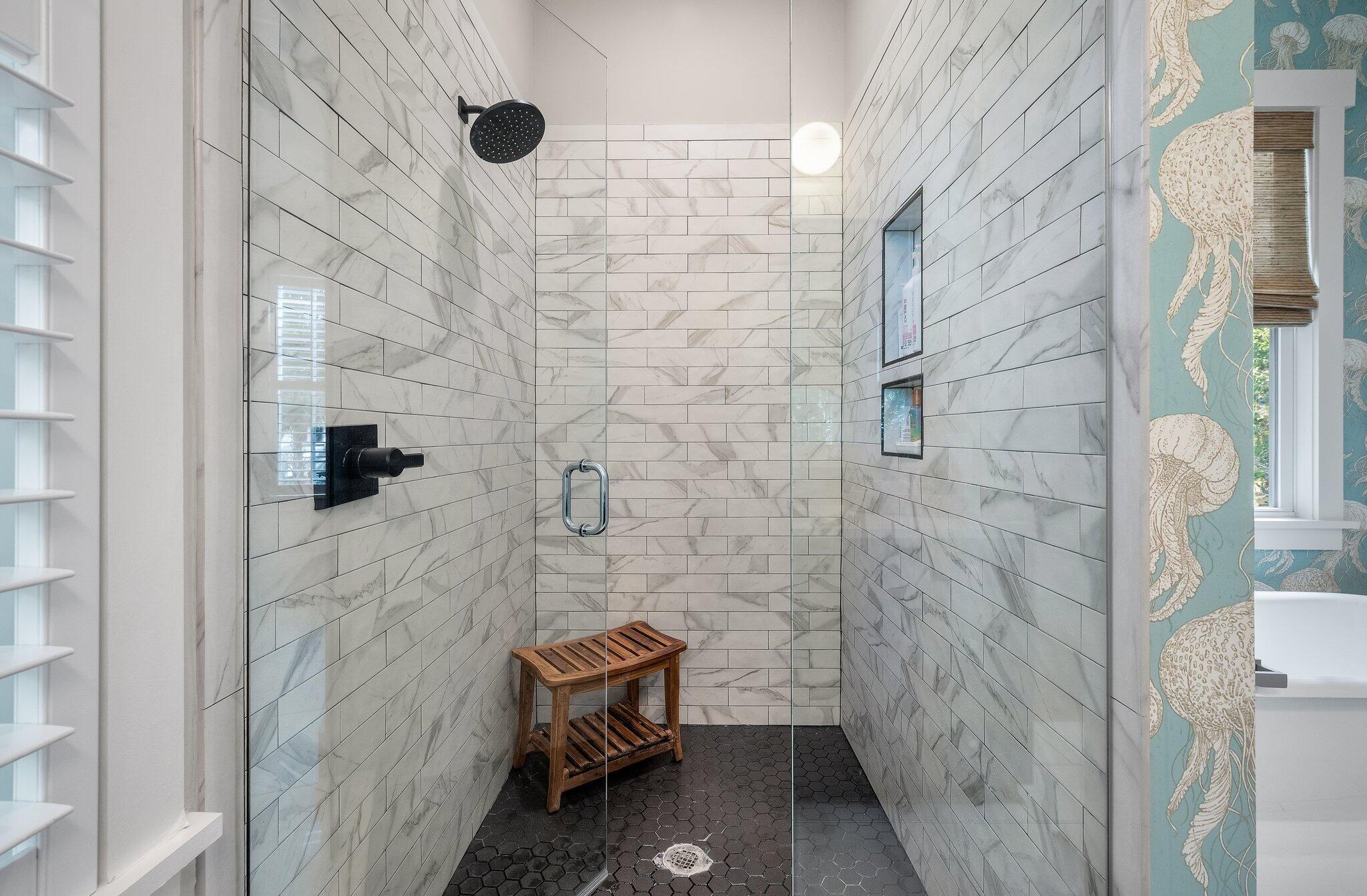 Walk-in shower with marble-look tile, a wooden bench, and black hexagonal flooring.