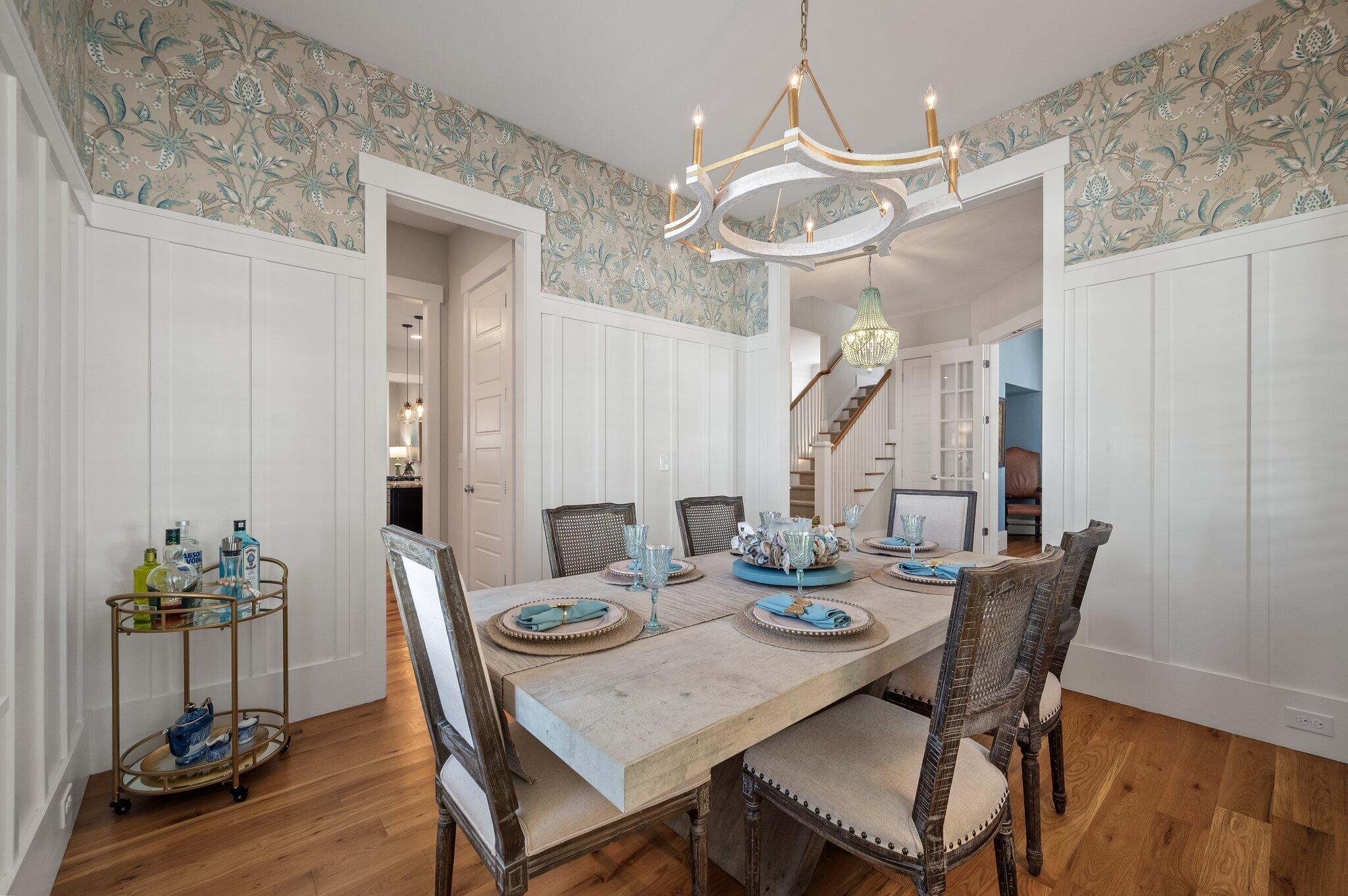 Staged dining room with a set table, patterned wallpaper, white wainscoting, and sculptural chandelier.