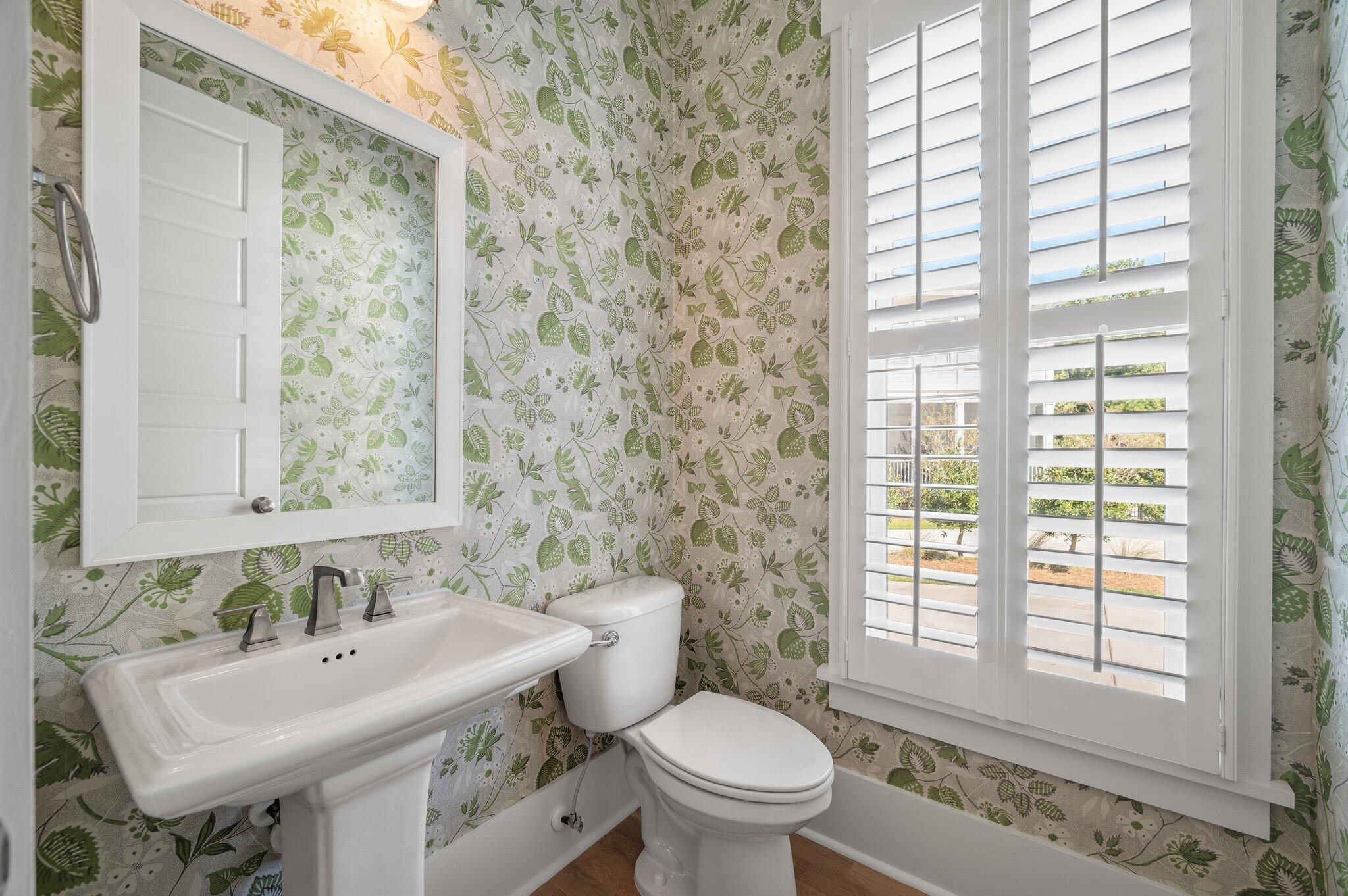 Bathroom with leafy floral wallpaper, pedestal sink, toilet, and plantation shutters.