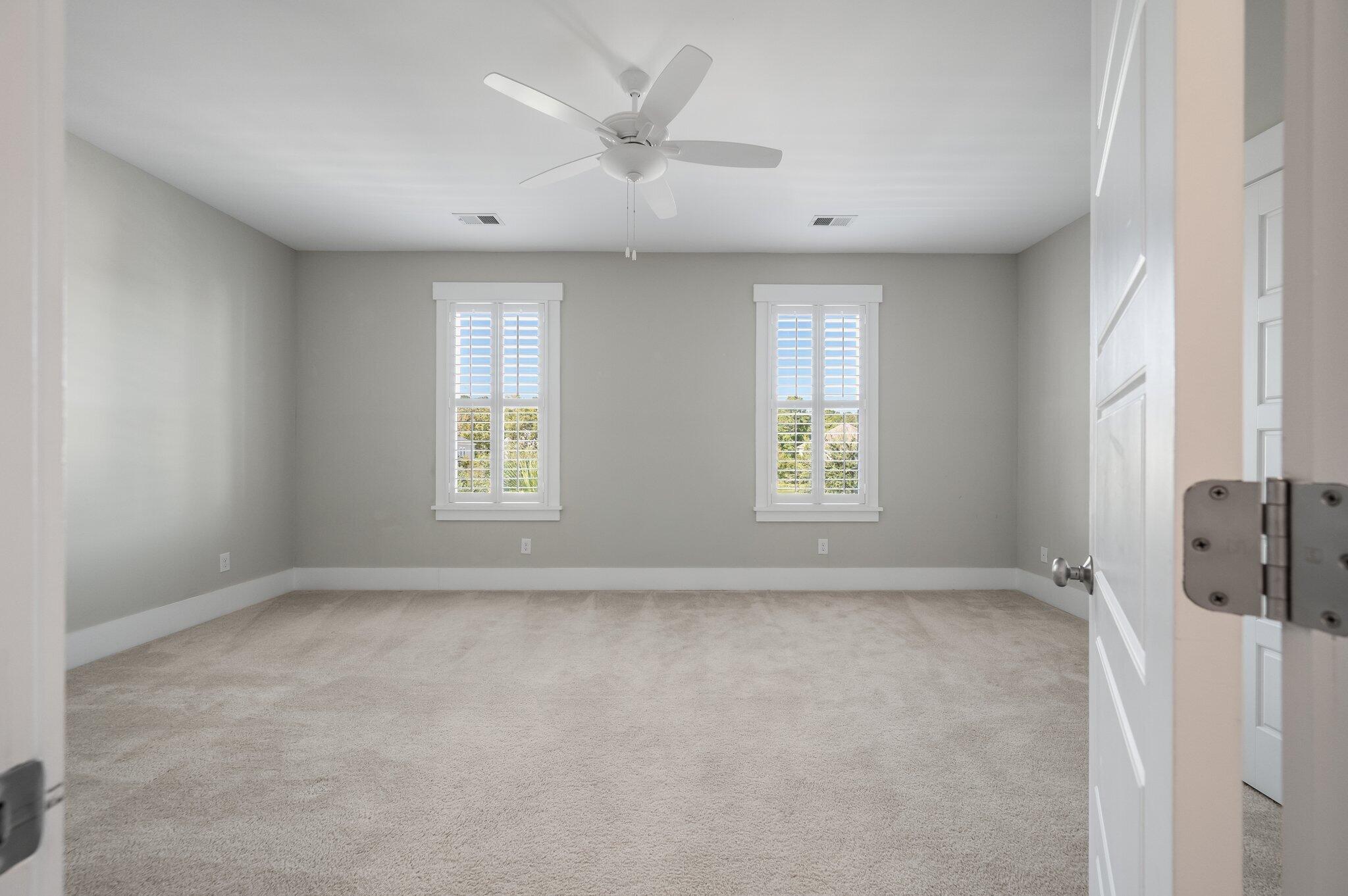 Empty carpeted bedroom with two shuttered windows and a ceiling fan.