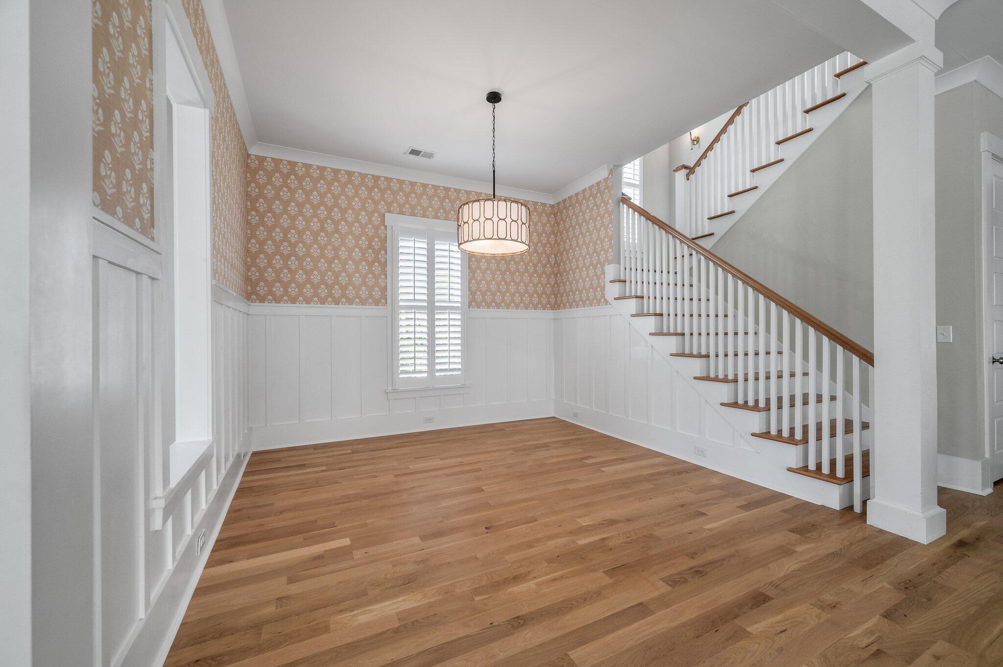 Dining room with patterned wallpaper, white wainscoting, hardwood floors, and an open staircase.