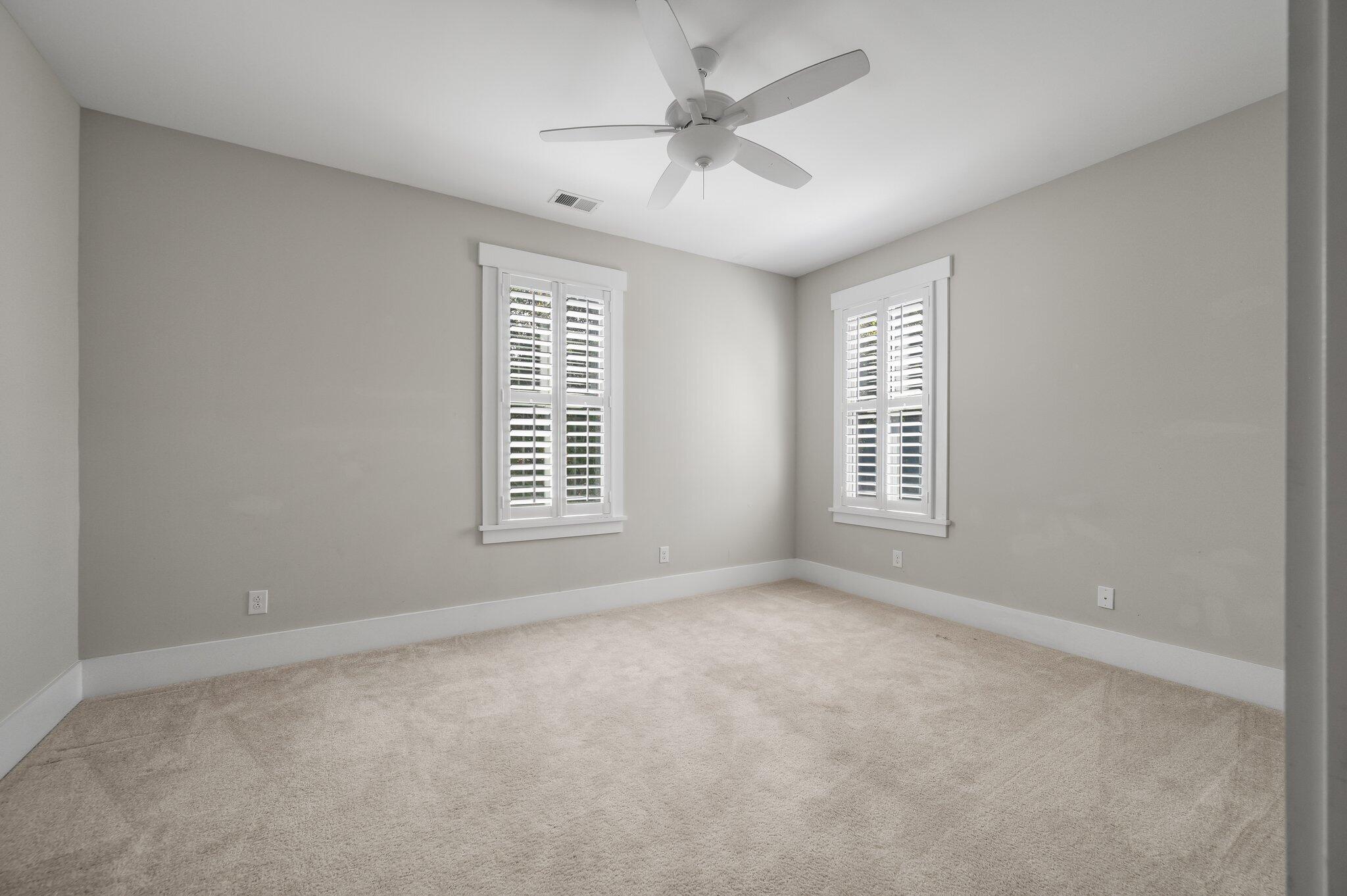 Empty carpeted room with two shuttered windows and a ceiling fan.