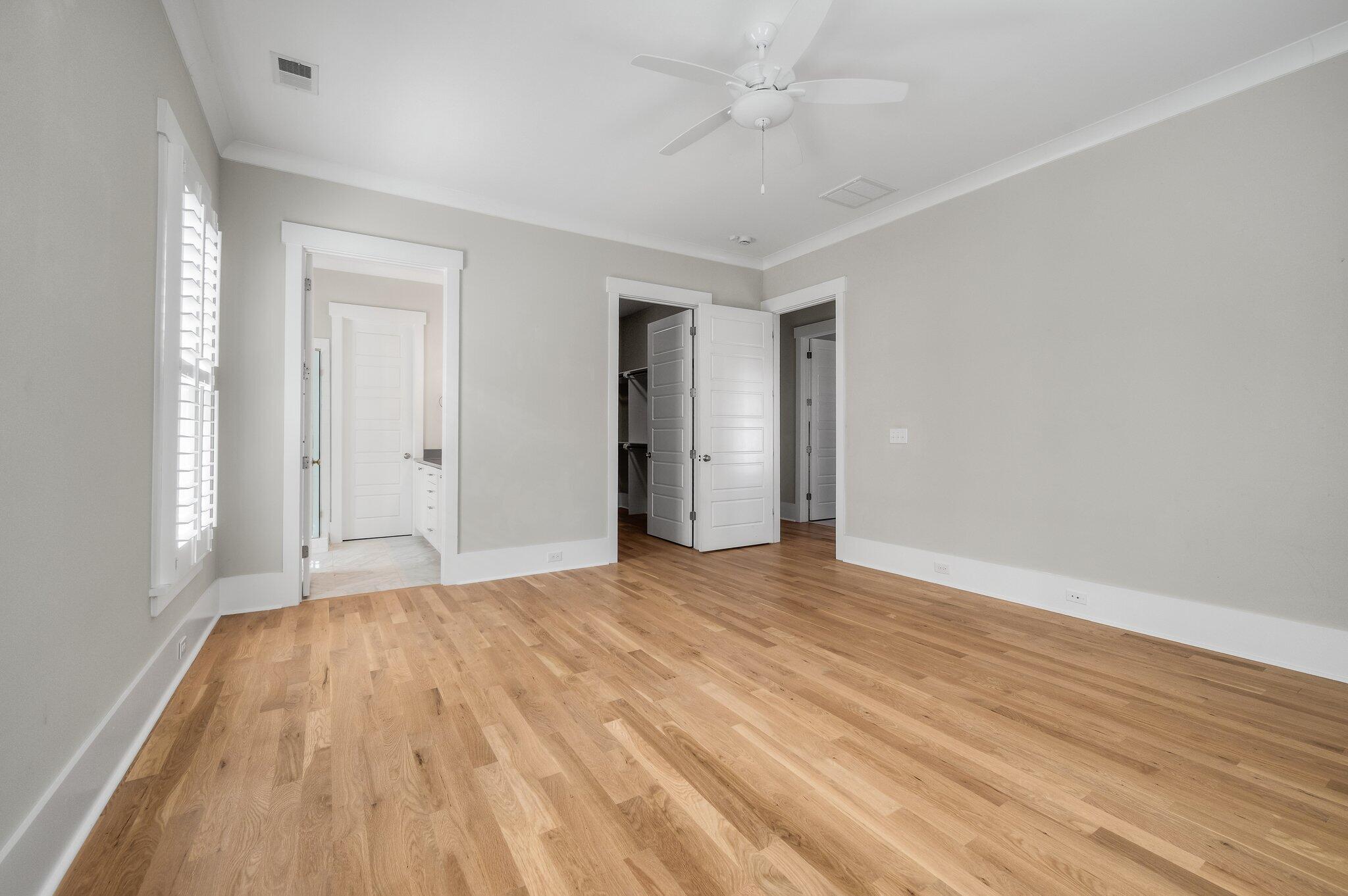 Empty bedroom with hardwood floors, plantation shutters, and doors to a bathroom and closet.