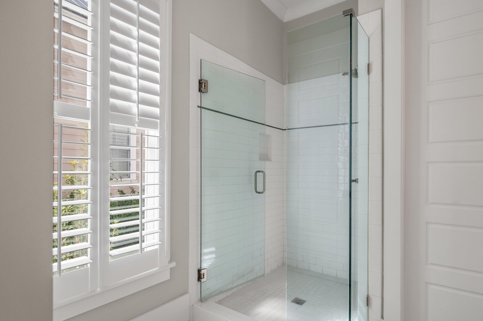 Walk-in shower with clear glass enclosure, white tile, and plantation-shuttered window.