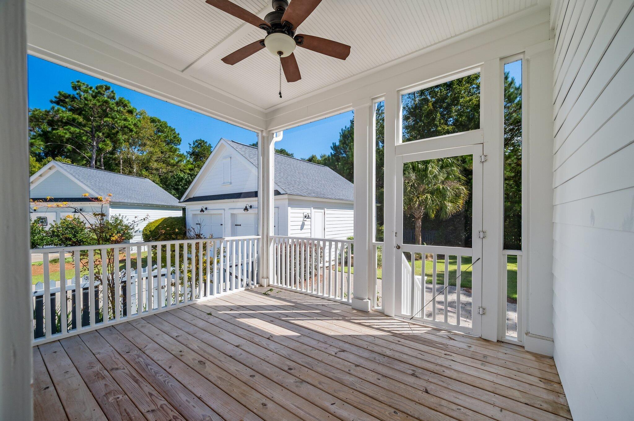 Covered porch with a ceiling fan, wood-plank floor, and white railings overlooking trees and neighboring homes.
