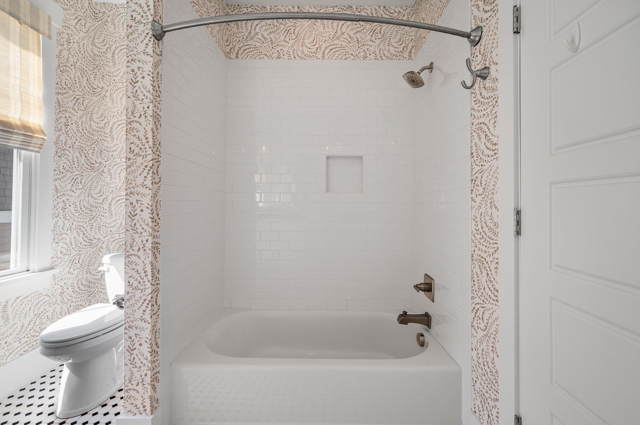 Bathroom with a white subway-tile tub and shower, patterned curtain, and toilet beside a window.